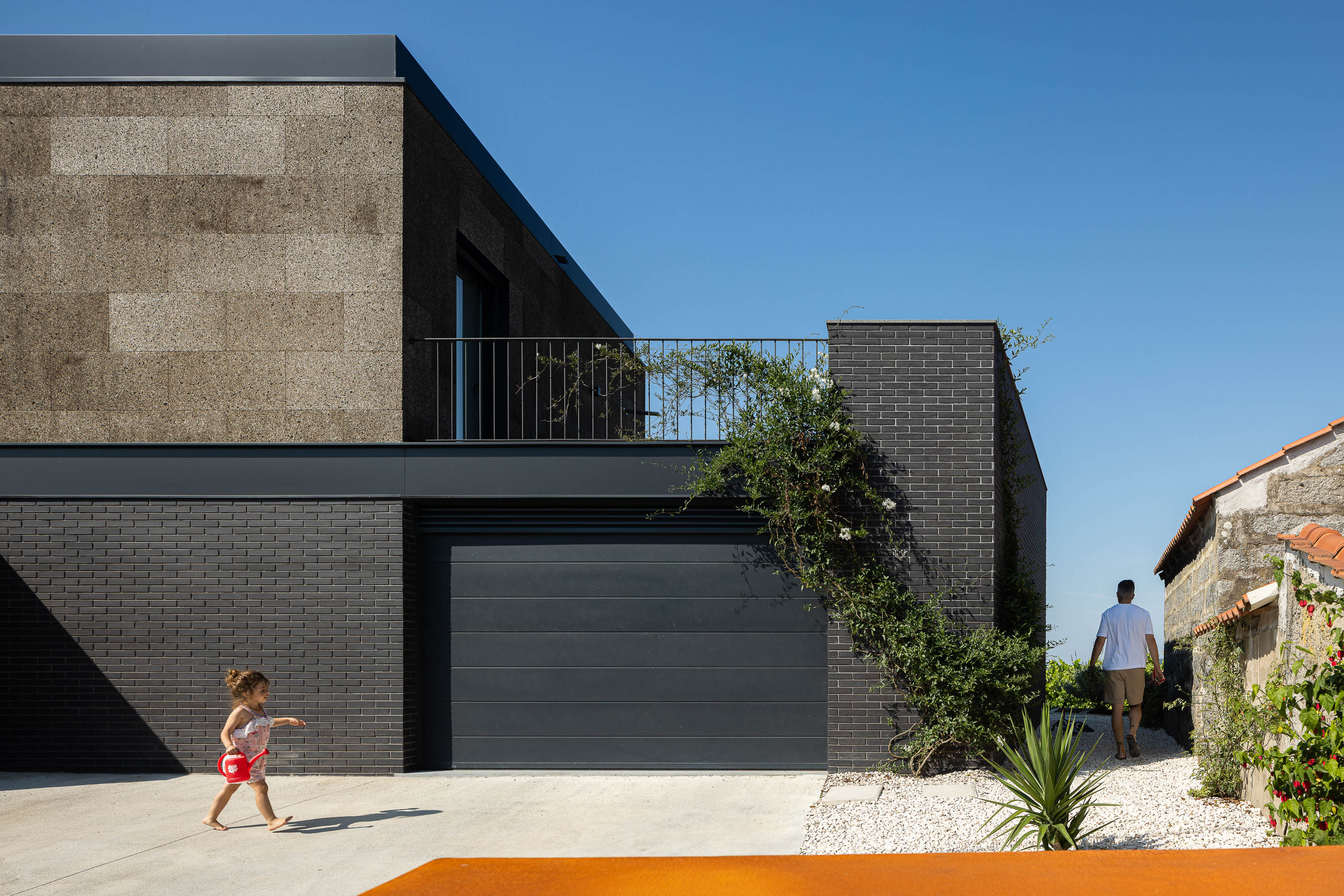 Exterior: Vista lateral da casa com garagem, revestimento de cortiça e tijolo preto, com vegetação. — Casa do Sobreiro arquitetura Skemba - Arquitectura e Engenharia em Amarante