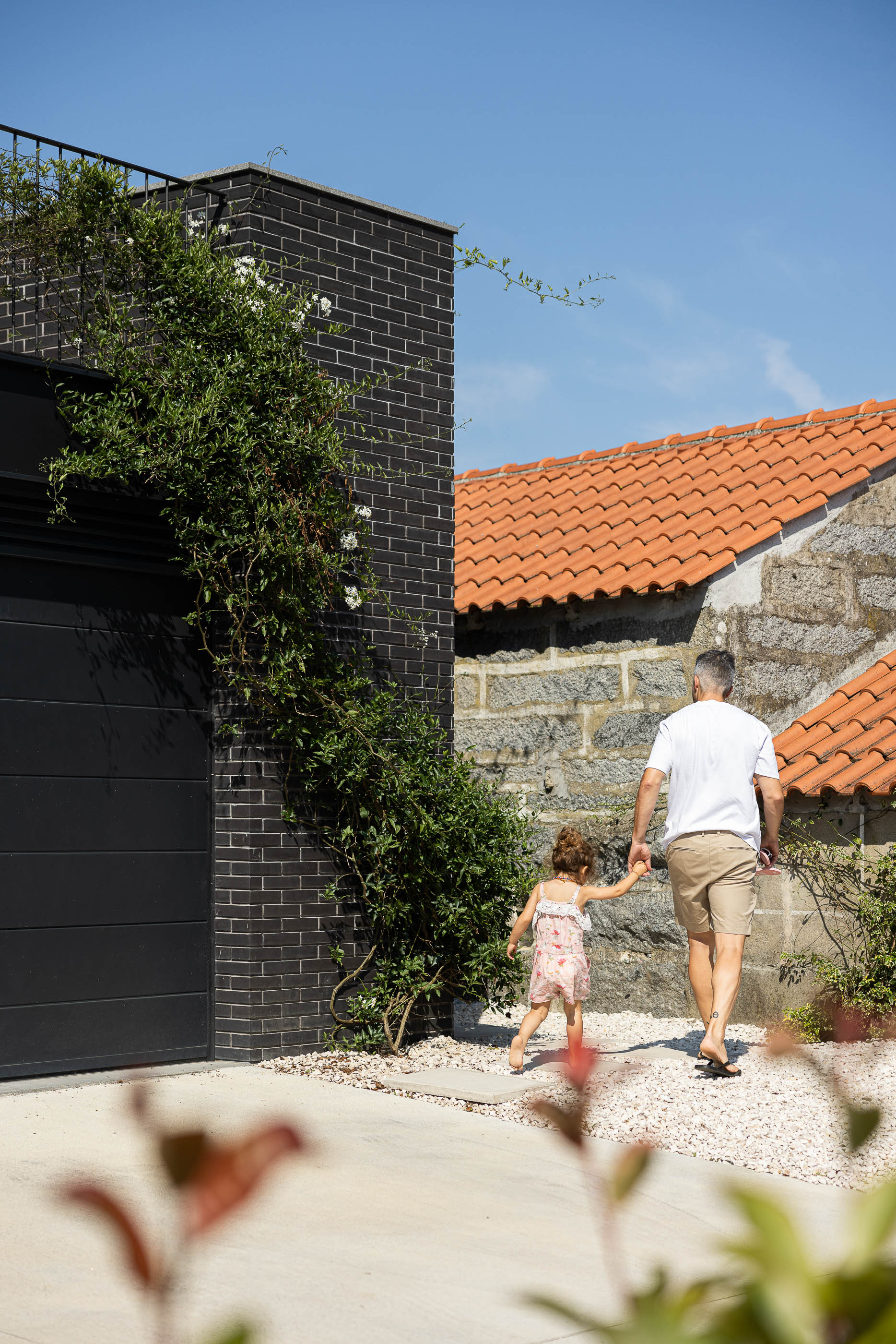 Exterior: Homem e criança caminham junto à garagem da casa com revestimento de tijolo preto e vegetação. — Casa do Sobreiro arquitetura Skemba - Arquitectura e Engenharia em Amarante