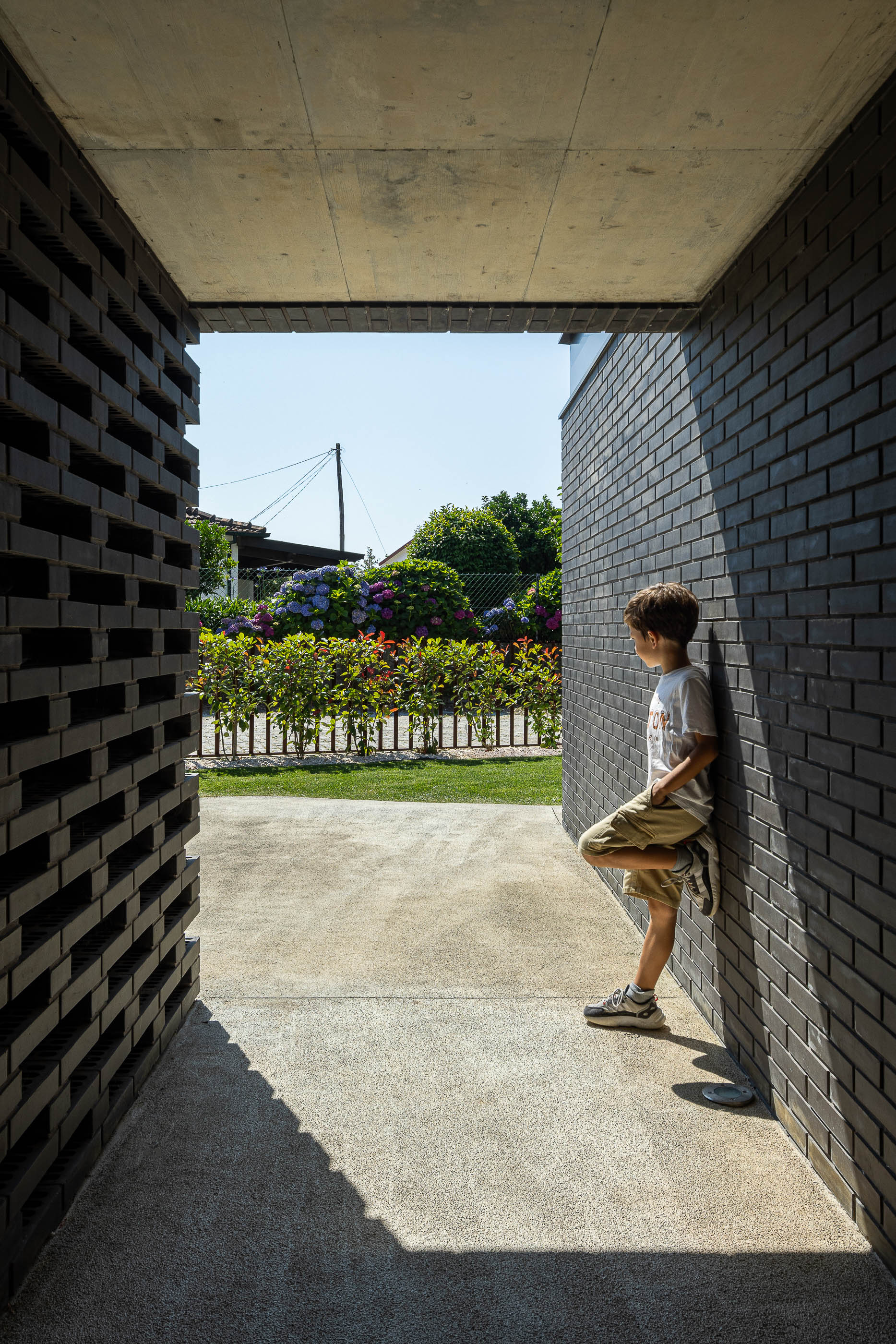 Entrada: Criança apoiada na parede de tijolo preto com elementos vazados, com vista para o jardim. — Casa do Sobreiro arquitetura Skemba - Arquitectura e Engenharia em Amarante