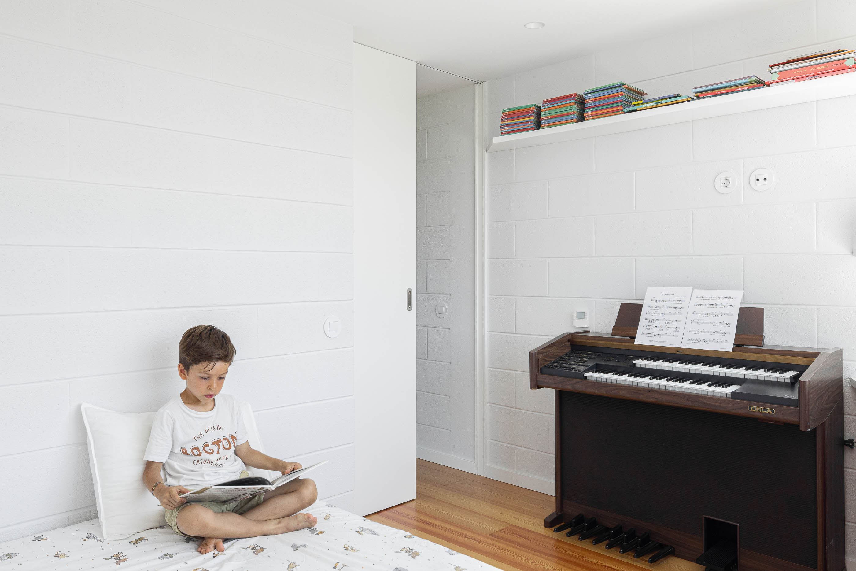 Quarto: Criança a ler num quarto com paredes brancas texturizadas, pavimento de madeira e um órgão. — Casa do Sobreiro arquitetura Skemba - Arquitectura e Engenharia em Amarante