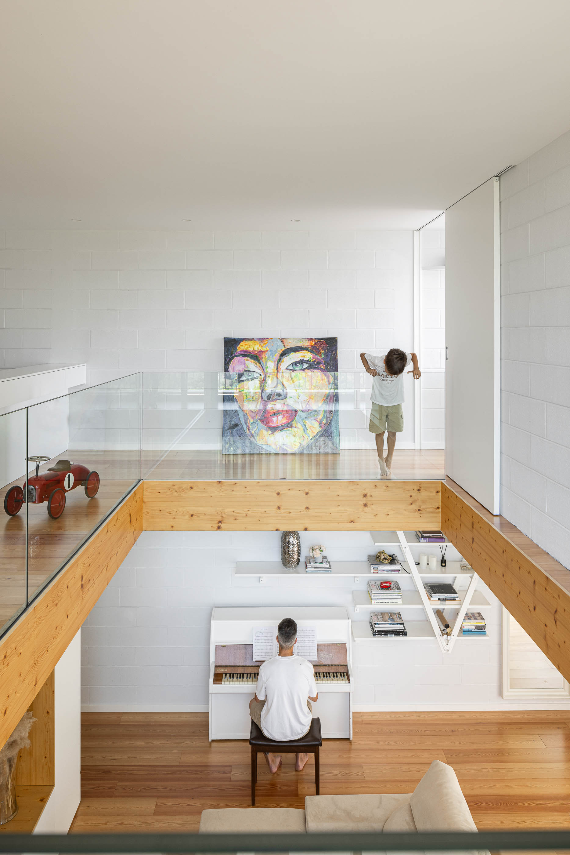 Mezzanine: Vista do espaço aberto com piano branco em baixo e criança no piso superior com guarda em vidro. — Casa do Sobreiro arquitetura Skemba - Arquitectura e Engenharia em Amarante