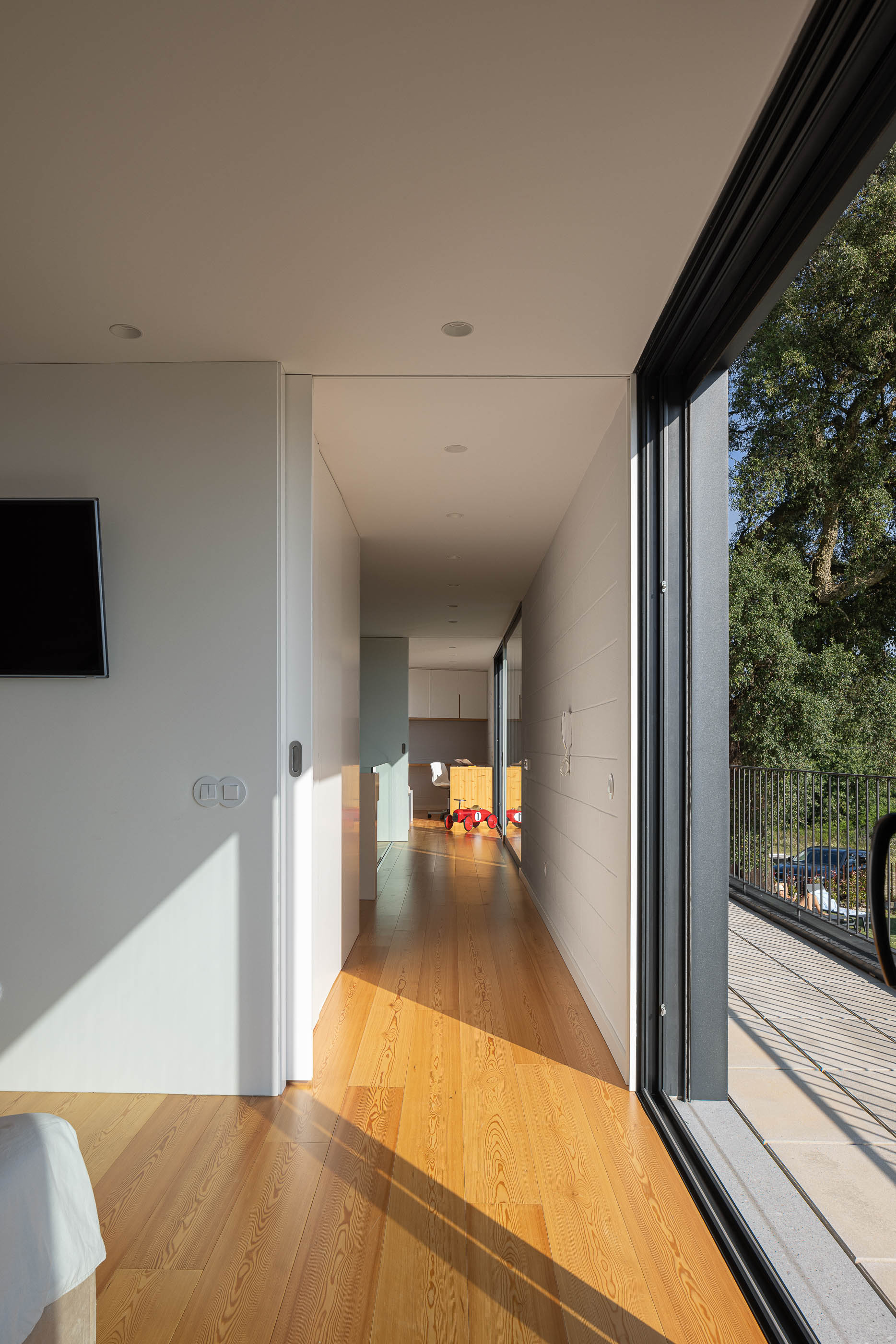 Corredor: Vista do corredor com pavimento de madeira, paredes brancas e acesso à varanda com vista para o jardim. — Casa do Sobreiro arquitetura Skemba - Arquitectura e Engenharia em Amarante