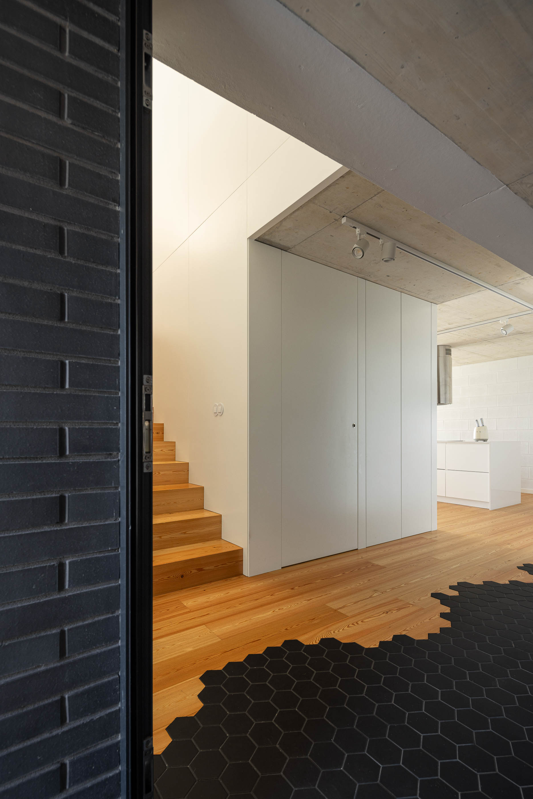 Entrada: Vista da entrada com pavimento de mosaico preto hexagonal, escadas de madeira e cozinha ao fundo. — Casa do Sobreiro arquitetura Skemba - Arquitectura e Engenharia em Amarante