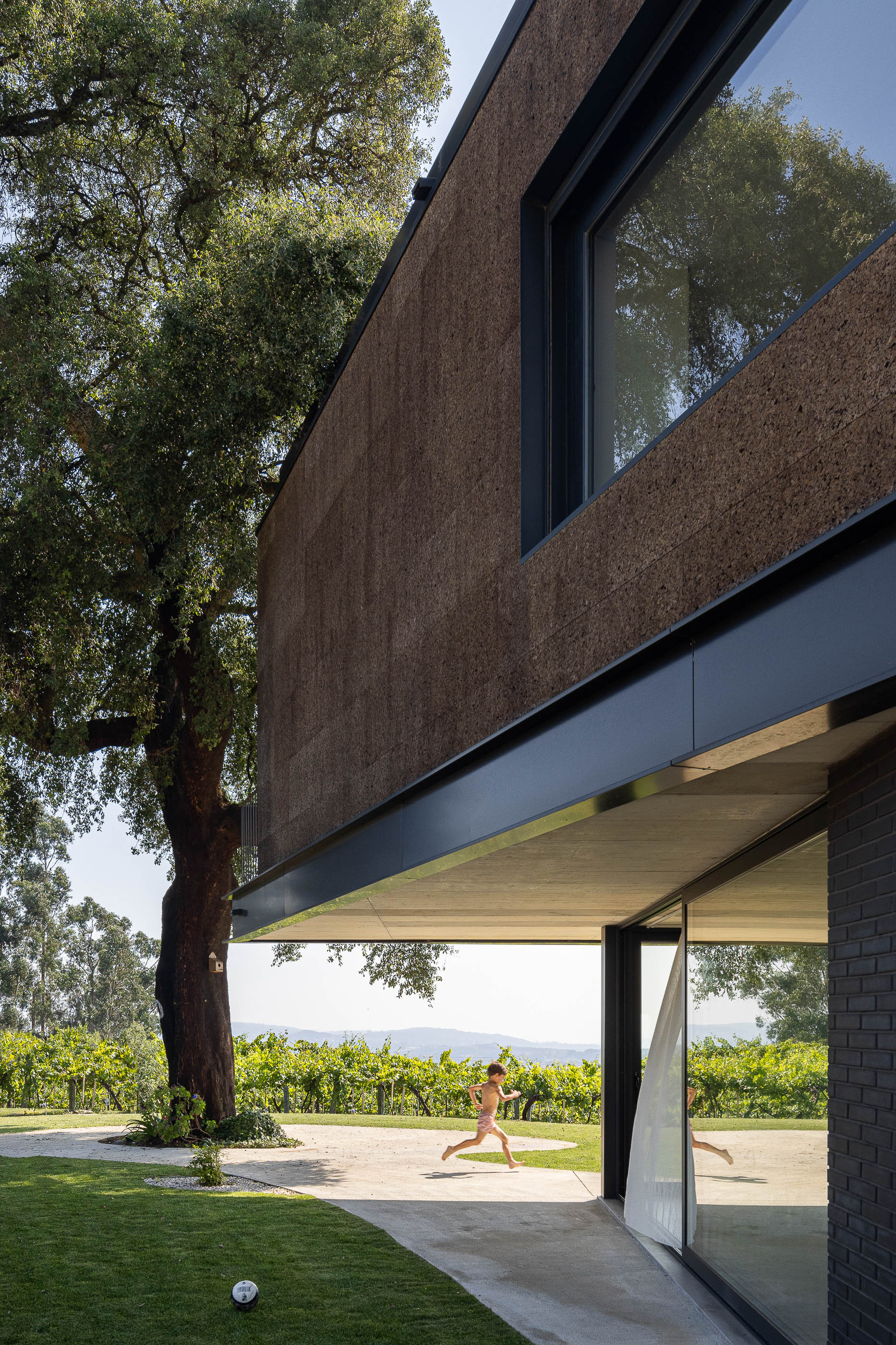 Exterior: Criança a brincar na varanda com guarda-corpo metálico, com um sobreiro ao lado. — Casa do Sobreiro arquitetura Skemba - Arquitectura e Engenharia em Amarante