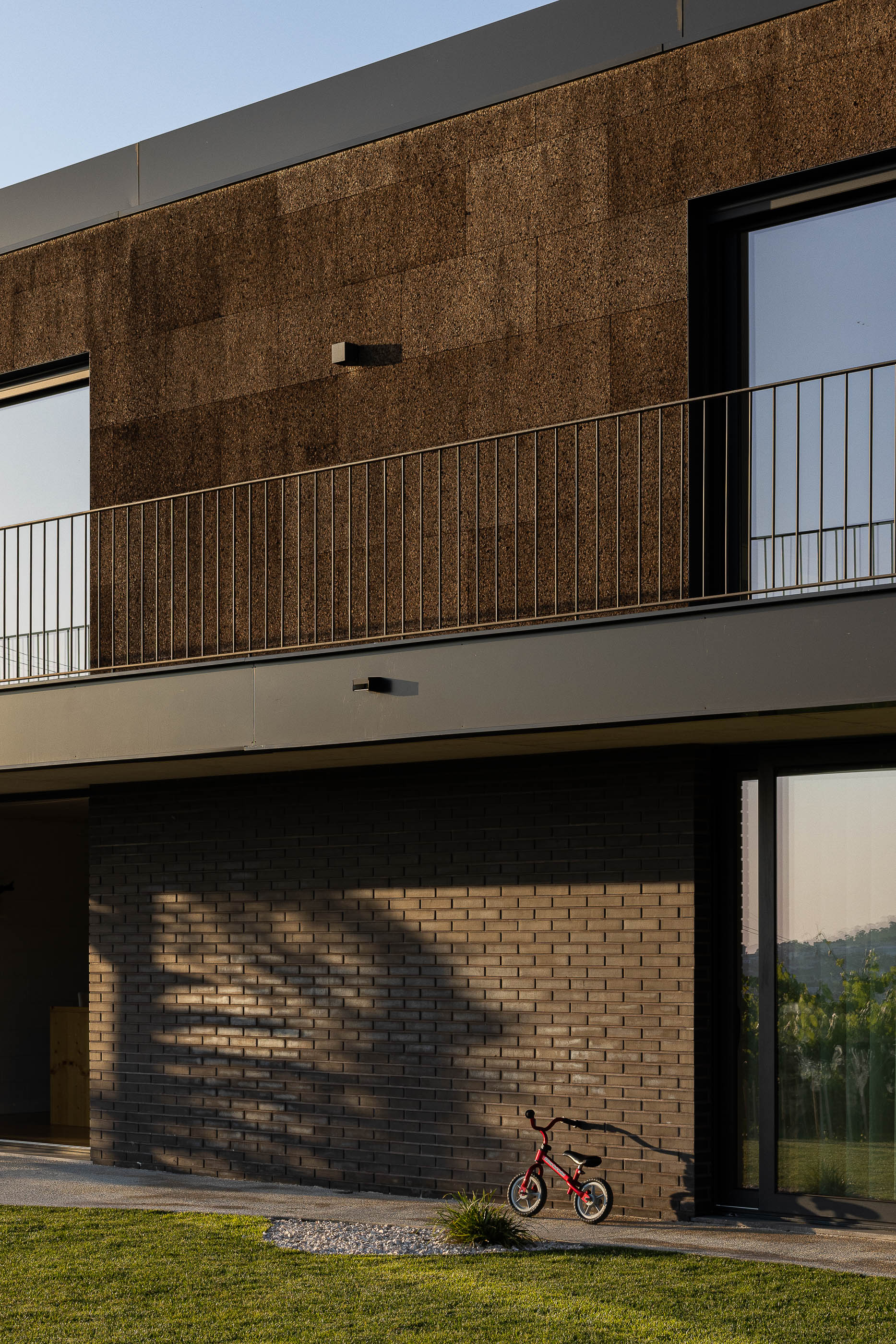 Exterior: Vista da casa com revestimento de cortiça e tijolo preto, com sobreiro e jardim com relvado. — Casa do Sobreiro arquitetura Skemba - Arquitectura e Engenharia em Amarante