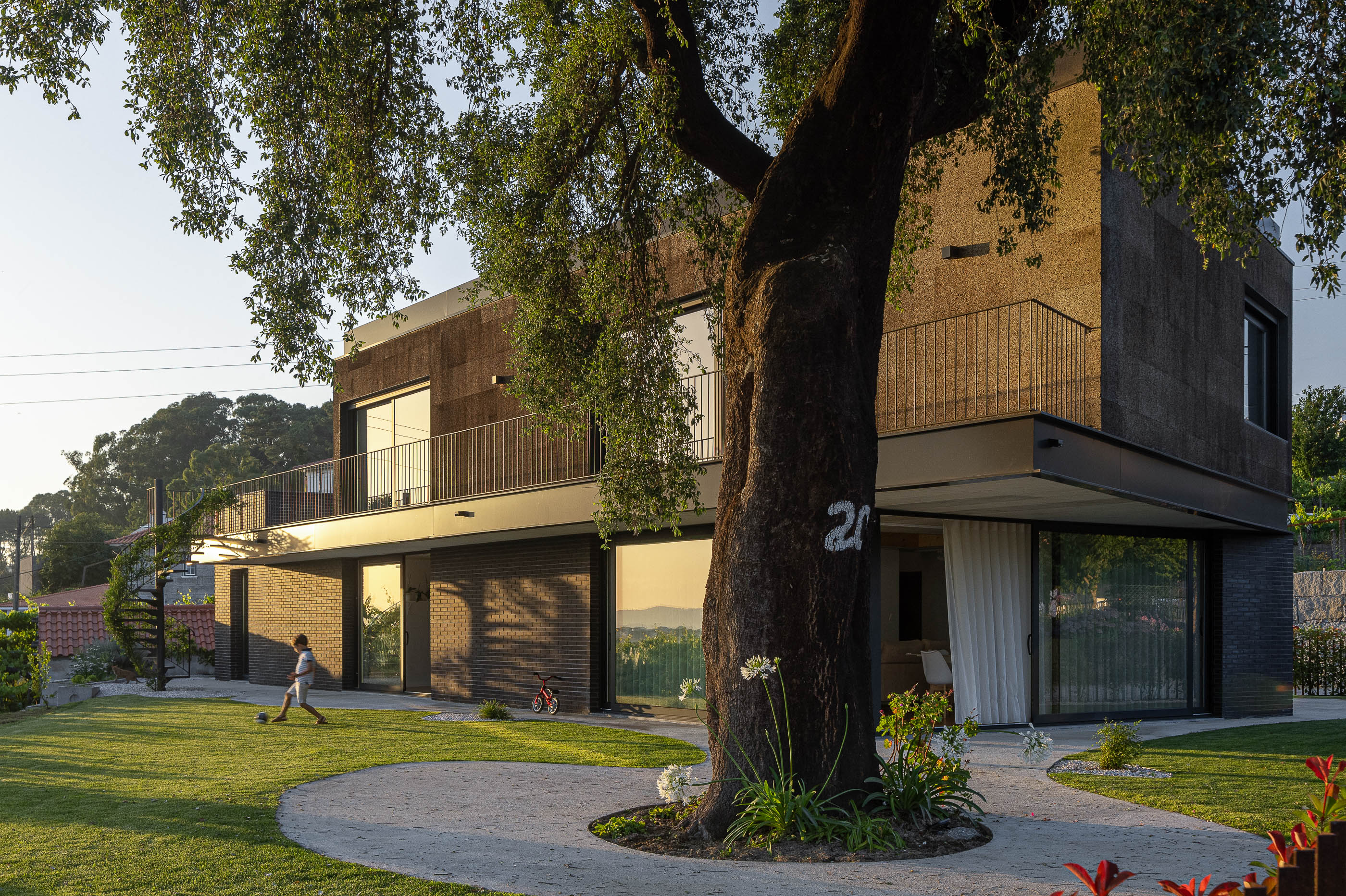 Exterior: Vista da fachada com revestimento de cortiça e tijolo preto, com sobreiro e vinhas ao fundo. — Casa do Sobreiro arquitetura Skemba - Arquitectura e Engenharia em Amarante