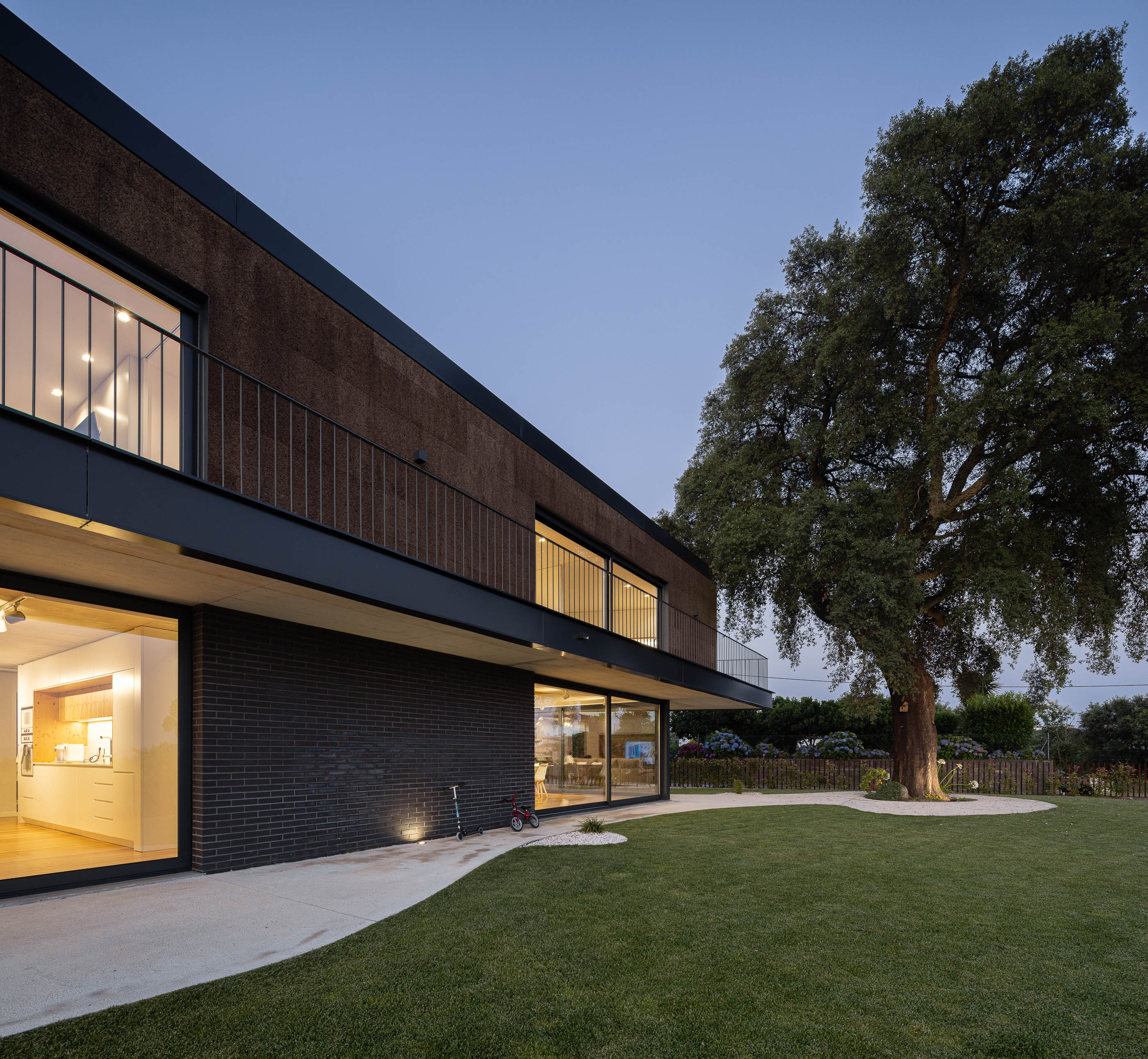 Exterior: Vista da casa iluminada ao entardecer, com revestimento de cortiça e tijolo preto, com sobreiro e vinhas ao fundo. — Casa do Sobreiro arquitetura Skemba - Arquitectura e Engenharia em Amarante