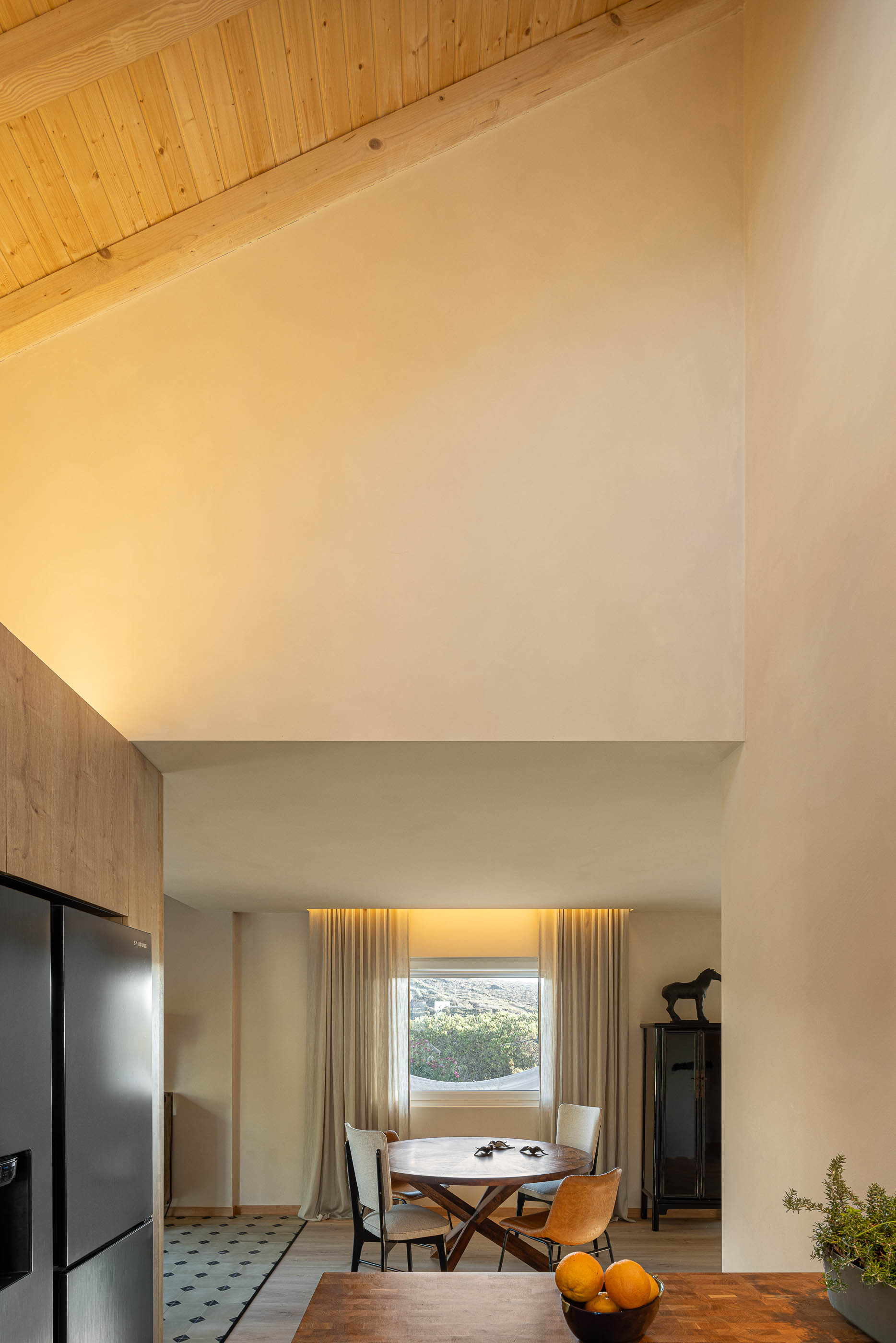 Cozinha/Sala de jantar: Vista vertical do espaço com teto alto em madeira, mesa redonda e armários em madeira clara. — Casa de Sintra arquitetura Tsou Arquitectos em Sintra