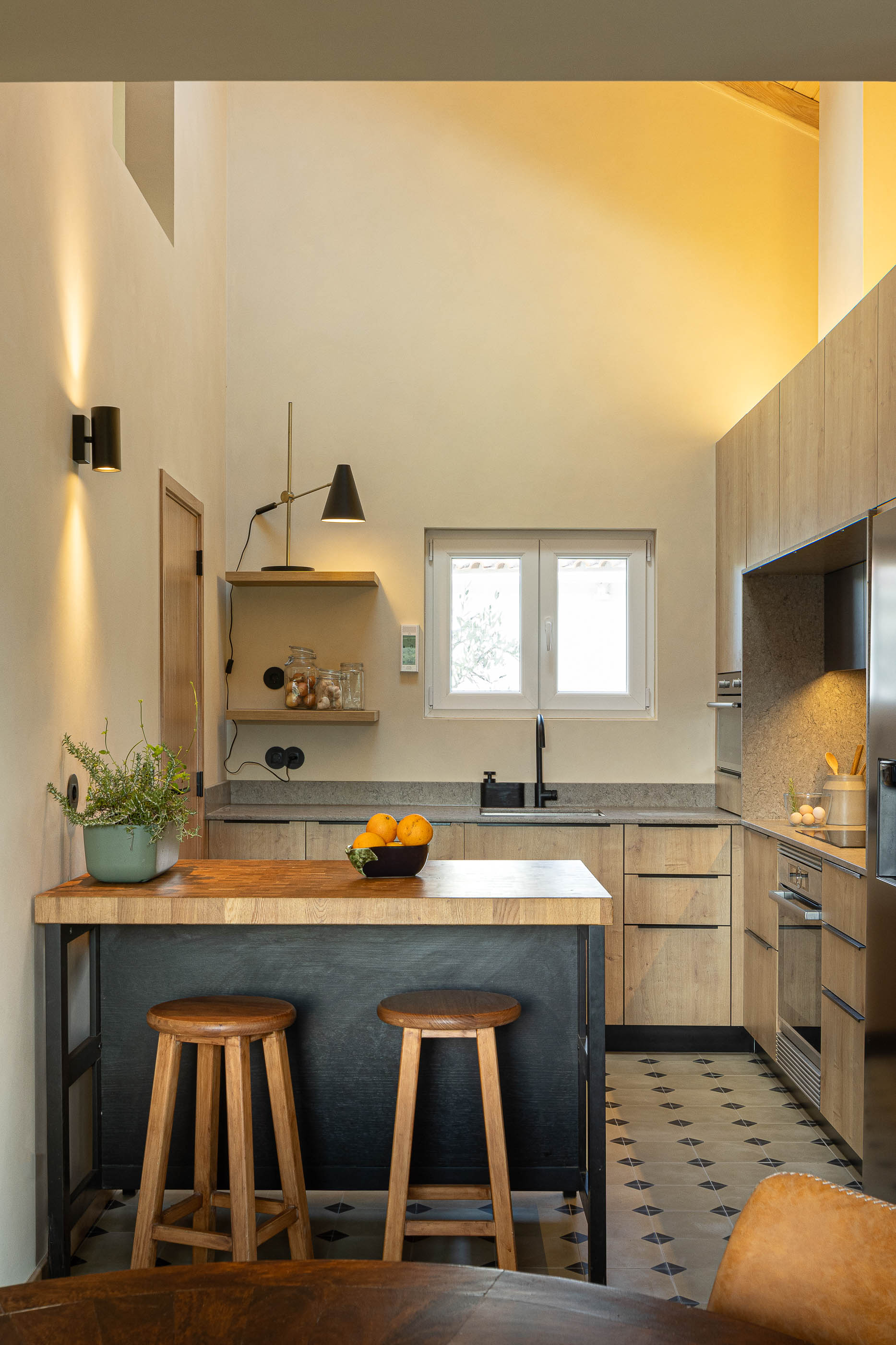Cozinha: Ilha central com bancada em madeira, cadeiras altas, armários em madeira clara e pavimento cerâmico. — Casa de Sintra arquitetura Tsou Arquitectos em Sintra