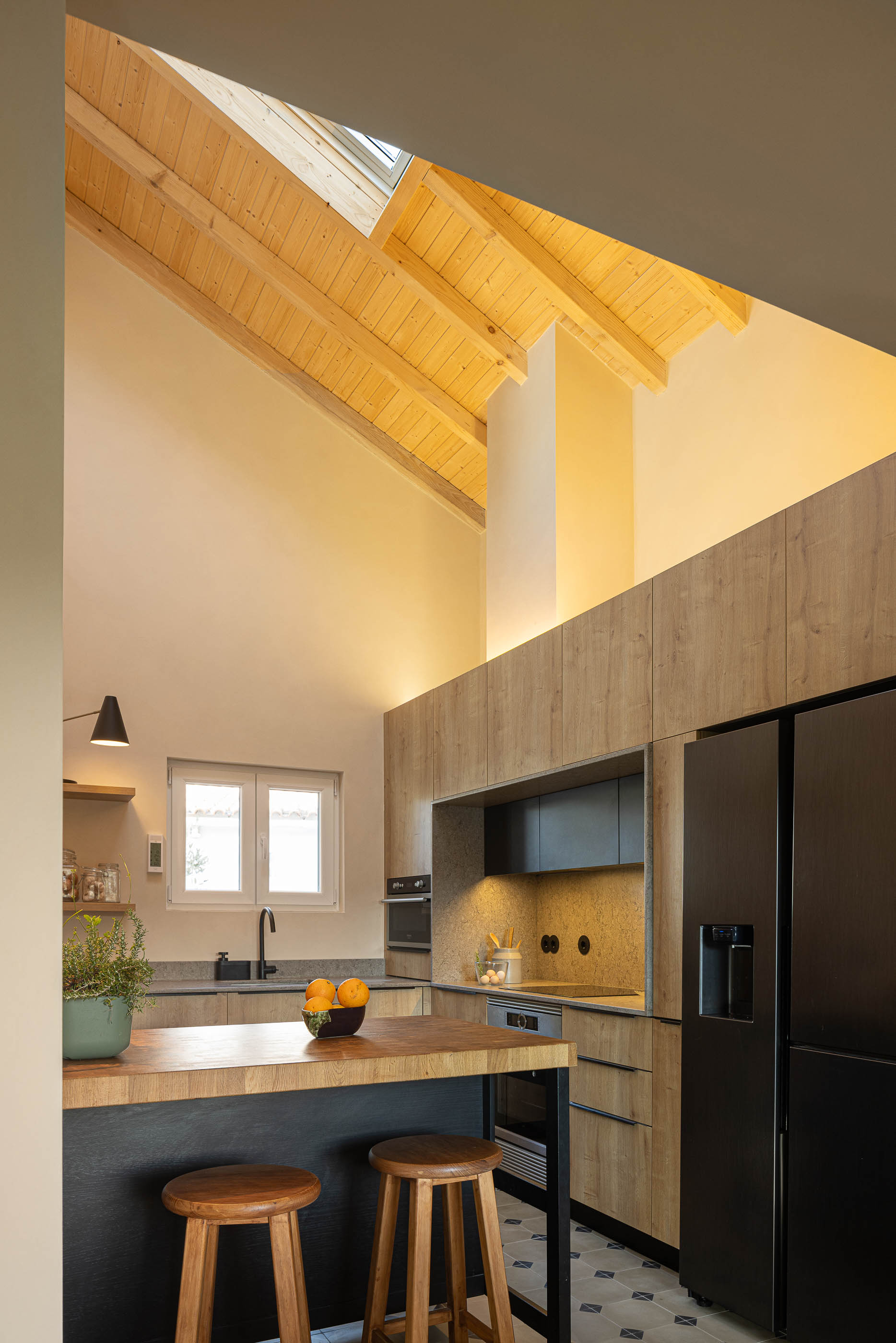 Cozinha: Vista geral da cozinha com teto alto em madeira, ilha central, armários em madeira clara e frigorífico preto. — Casa de Sintra arquitetura Tsou Arquitectos em Sintra