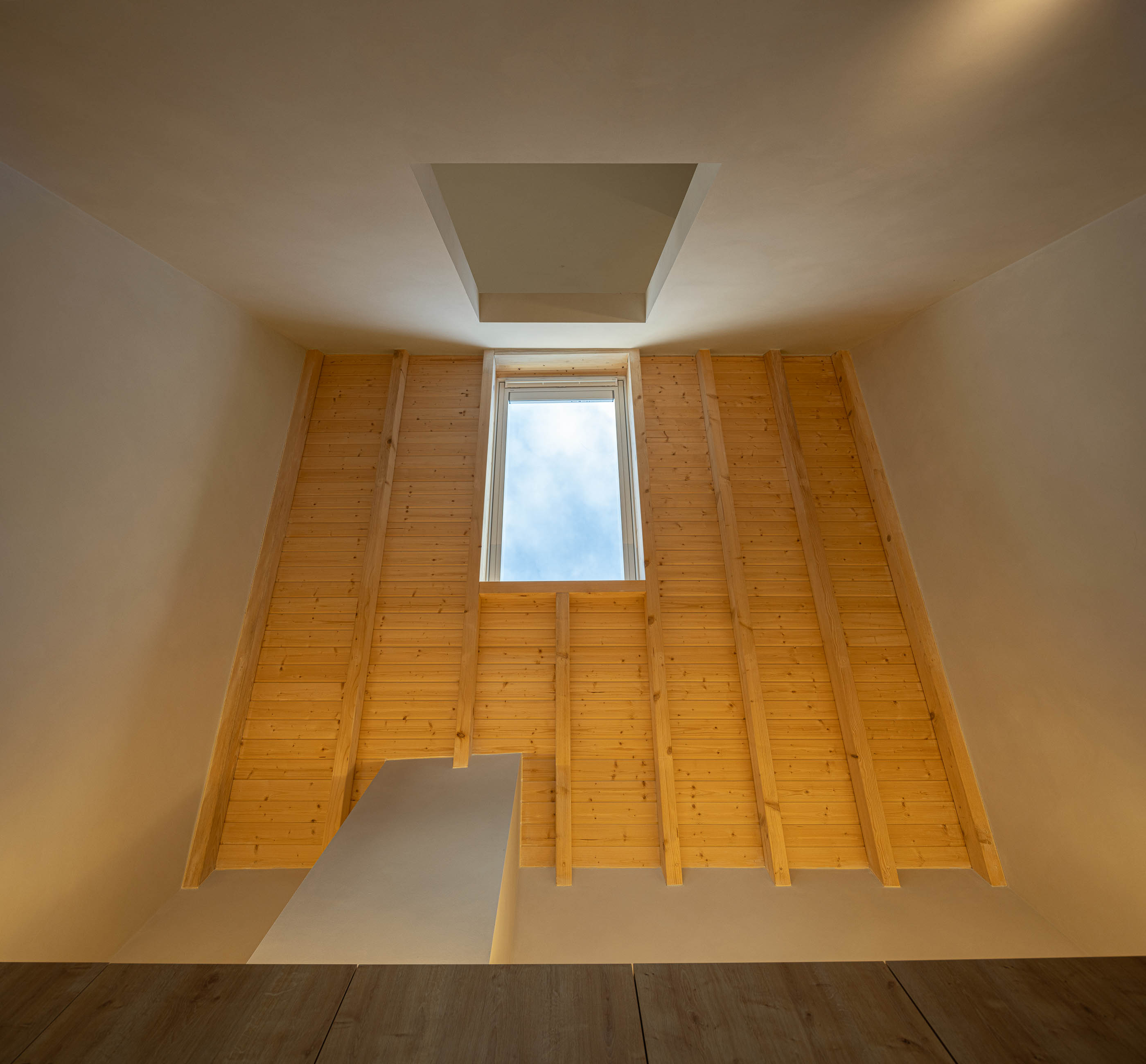 Sótão: Teto inclinado com janela e parede revestida a madeira clara. — Casa de Sintra arquitetura Tsou Arquitectos em Sintra