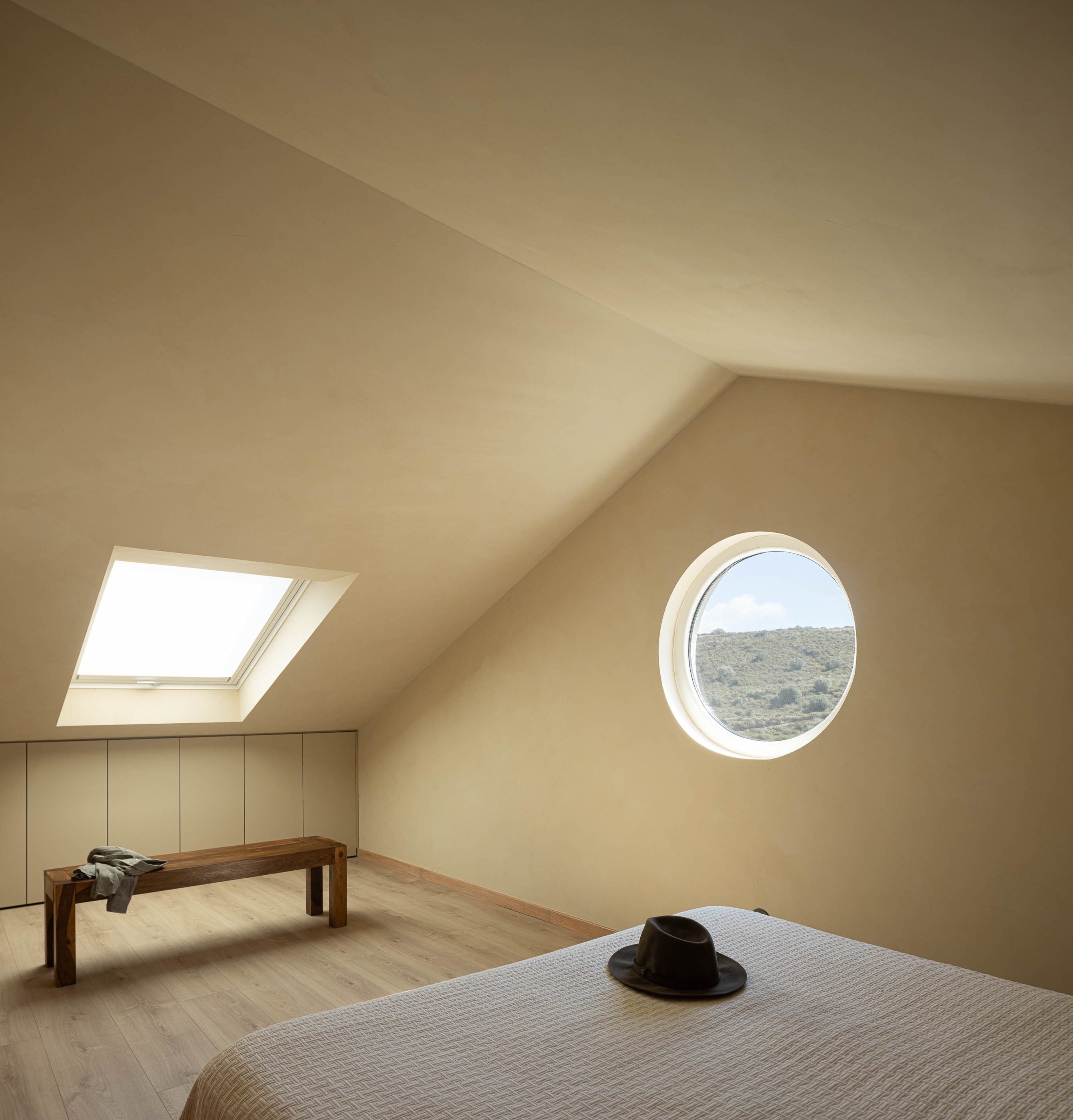 Quarto: Quarto no sótão com janela redonda, cama com colcha branca e pavimento em madeira clara. — Casa de Sintra arquitetura Tsou Arquitectos em Sintra