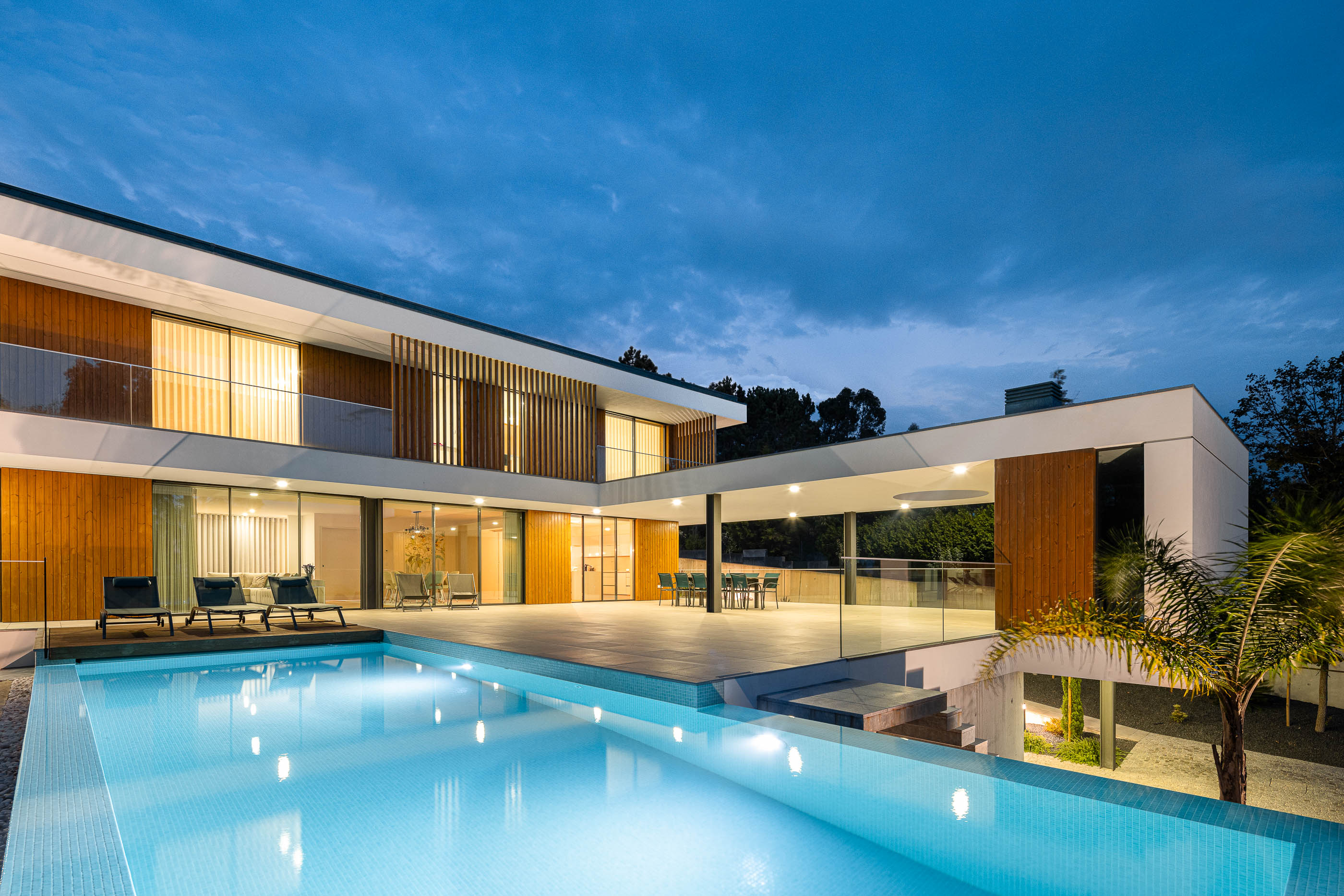Exterior: Vista noturna da casa com piscina iluminada, terraço e detalhes em madeira, arquitetura moderna e elegante. — Casa J.M.C. arquitetura Atelier d´Arquitectura Lopes da Costa em Santa Maria da Feira