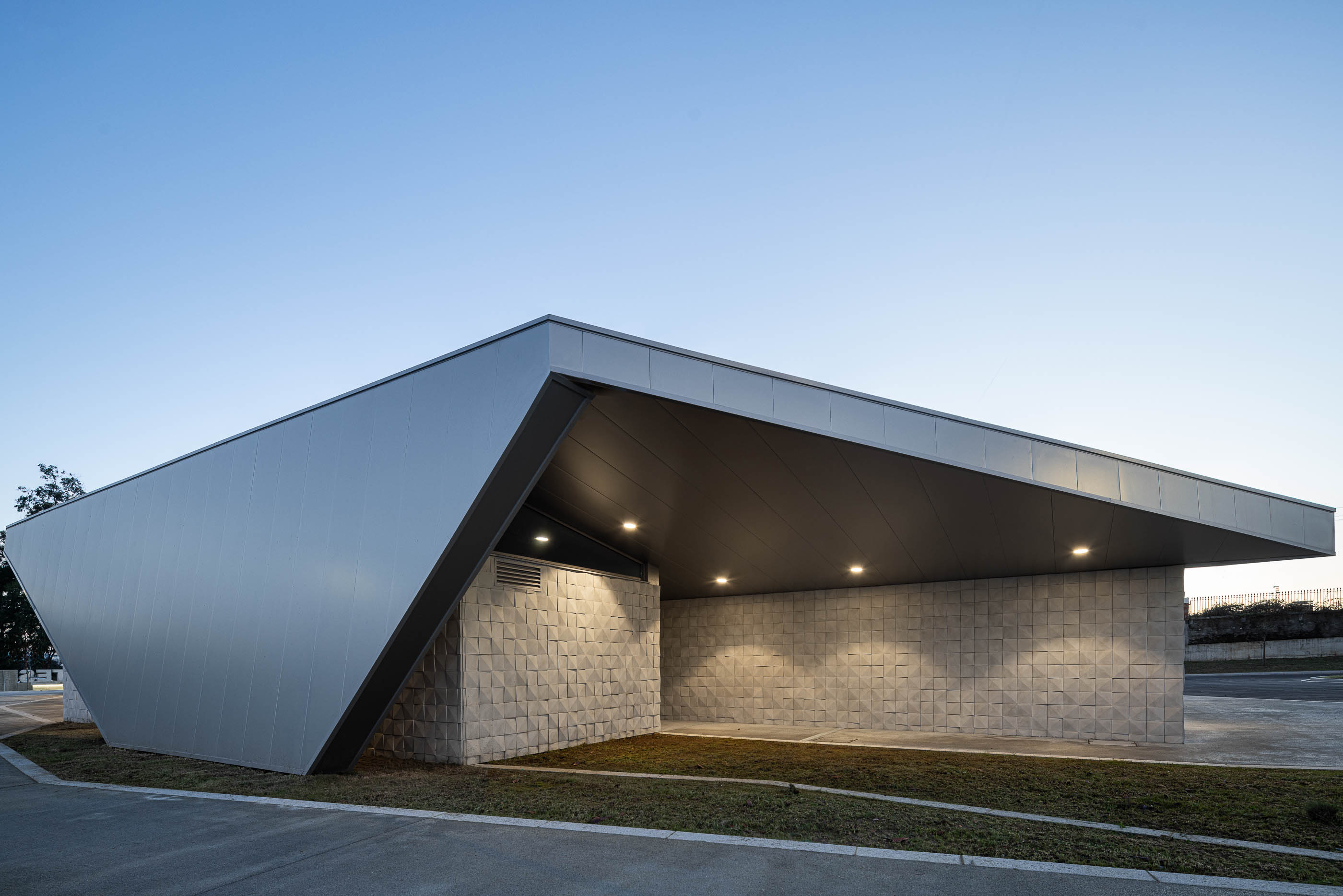 Exterior: Vista lateral do terminal com iluminação embutida, fachada texturada e estacionamento. — Interface de Transportes Lourosa-Fiães arquitetura Atelier d´Arquitectura Lopes da Costa em Santa Maria da Feira