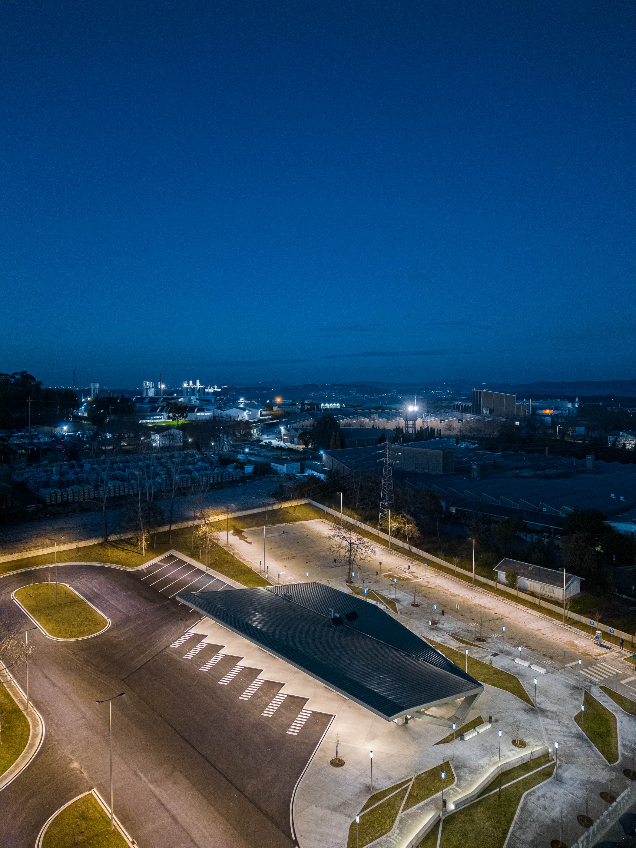 Vista aérea noturna: Terminal de transportes com estacionamento iluminado e paisagem urbana ao fundo. — Interface de Transportes Lourosa-Fiães arquitetura Atelier d´Arquitectura Lopes da Costa em Santa Maria da Feira