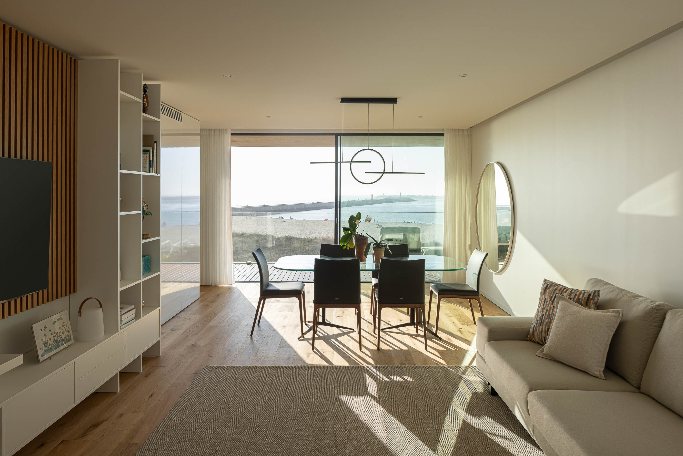 Sala de estar: Sala de estar com sofá, mesa de jantar, candeeiro suspenso e vista para a praia. — Edifício LUX arquitetura Mário Alves Arquitecto em Ílhavo