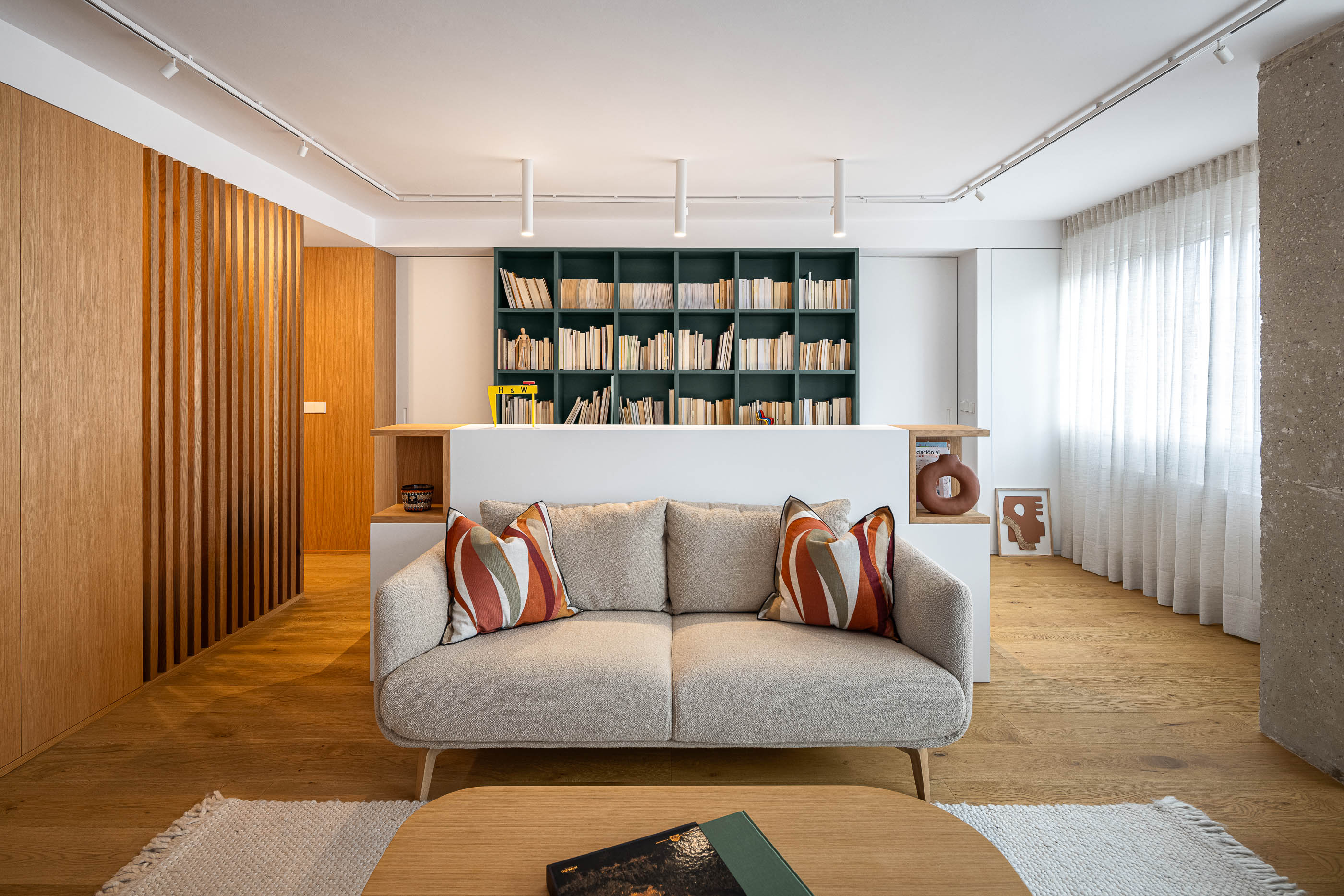 Sala de estar com sofá cinzento, almofadas coloridas e mesa de centro em madeira. Estante verde escura divide o espaço. Alcatifa clara. — Reforma Apartamento CV em Avilés arquitetura David Olmos Arquitectos em Avilés