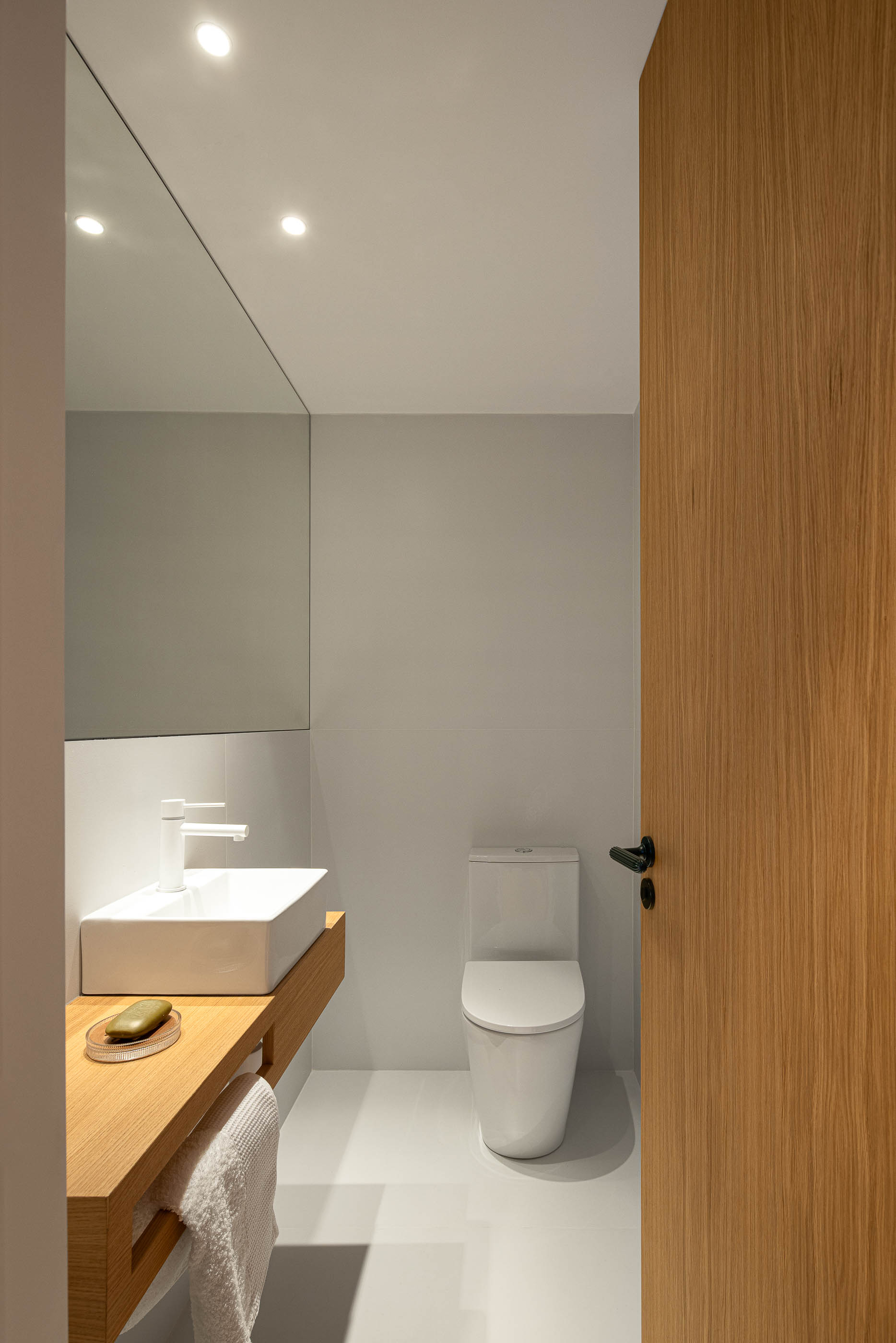 Casa de banho com lavatório branco sobre móvel de madeira clara, espelho e sanita. Pavimento branco e porta de madeira clara. — Reforma Apartamento CV em Avilés arquitetura David Olmos Arquitectos em Avilés