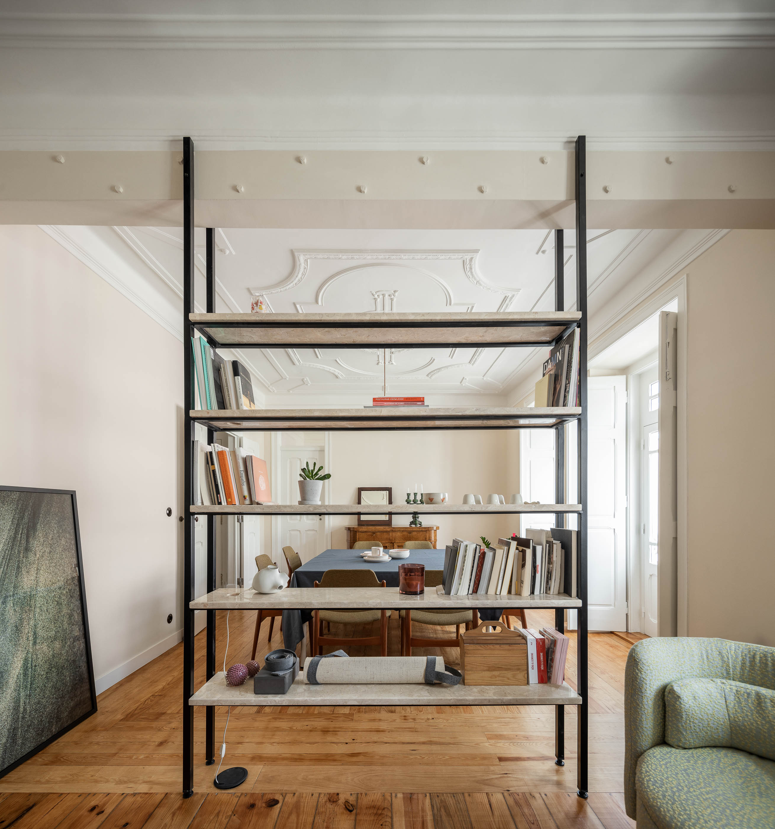 Sala: Estante metálica com prateleiras em pedra, pavimento em madeira, tecto trabalhado, mesa de jantar ao fundo. — Apartamento da Penha arquitetura Paulo Moreira Architectures em Lisboa