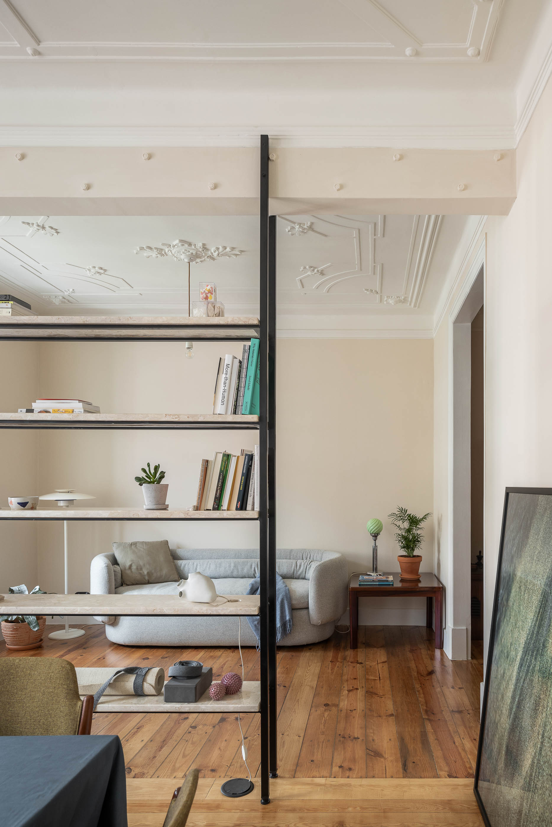 Sala: Estante metálica com prateleiras em pedra, pavimento em madeira, tecto trabalhado, sofá cinzento ao fundo. — Apartamento da Penha arquitetura Paulo Moreira Architectures em Lisboa