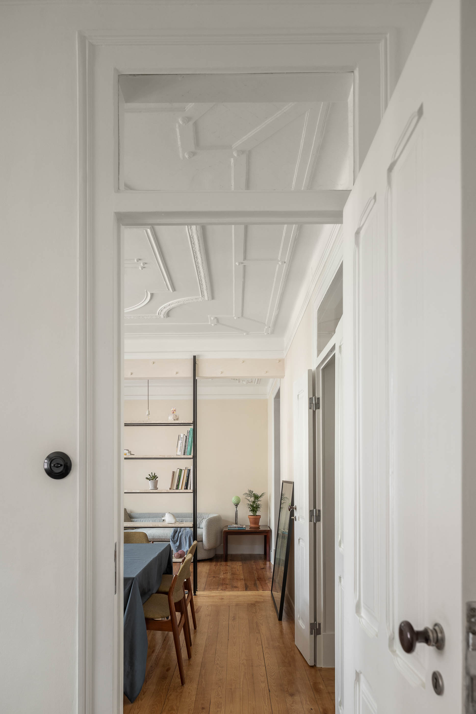 Corredor: Porta aberta para sala com estante, pavimento em madeira, tecto trabalhado, interruptor antigo na parede. — Apartamento da Penha arquitetura Paulo Moreira Architectures em Lisboa