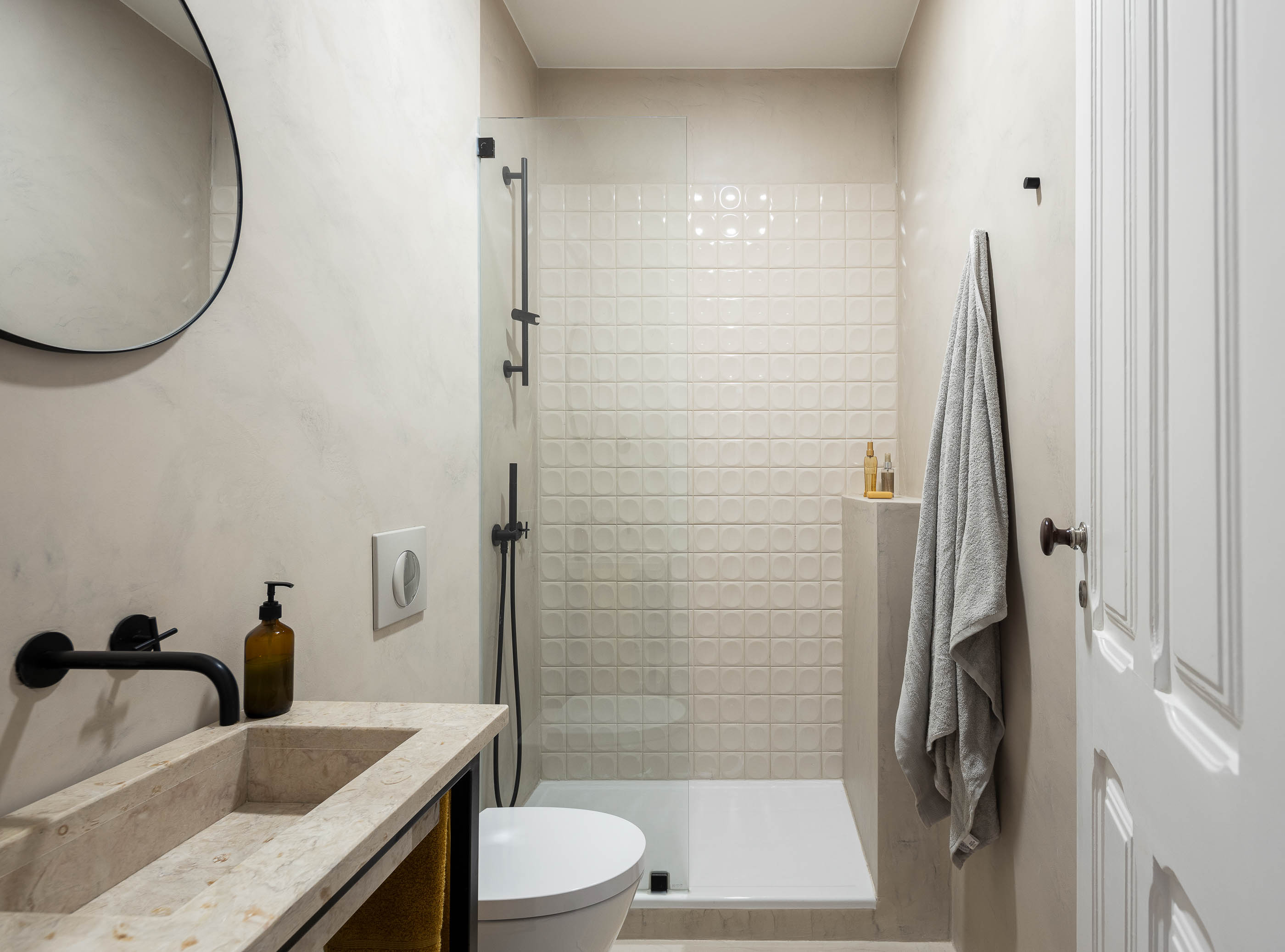Casa de banho: Lavatório em pedra com torneira preta, espelho redondo, duche com mosaico quadrado. — Apartamento da Penha arquitetura Paulo Moreira Architectures em Lisboa