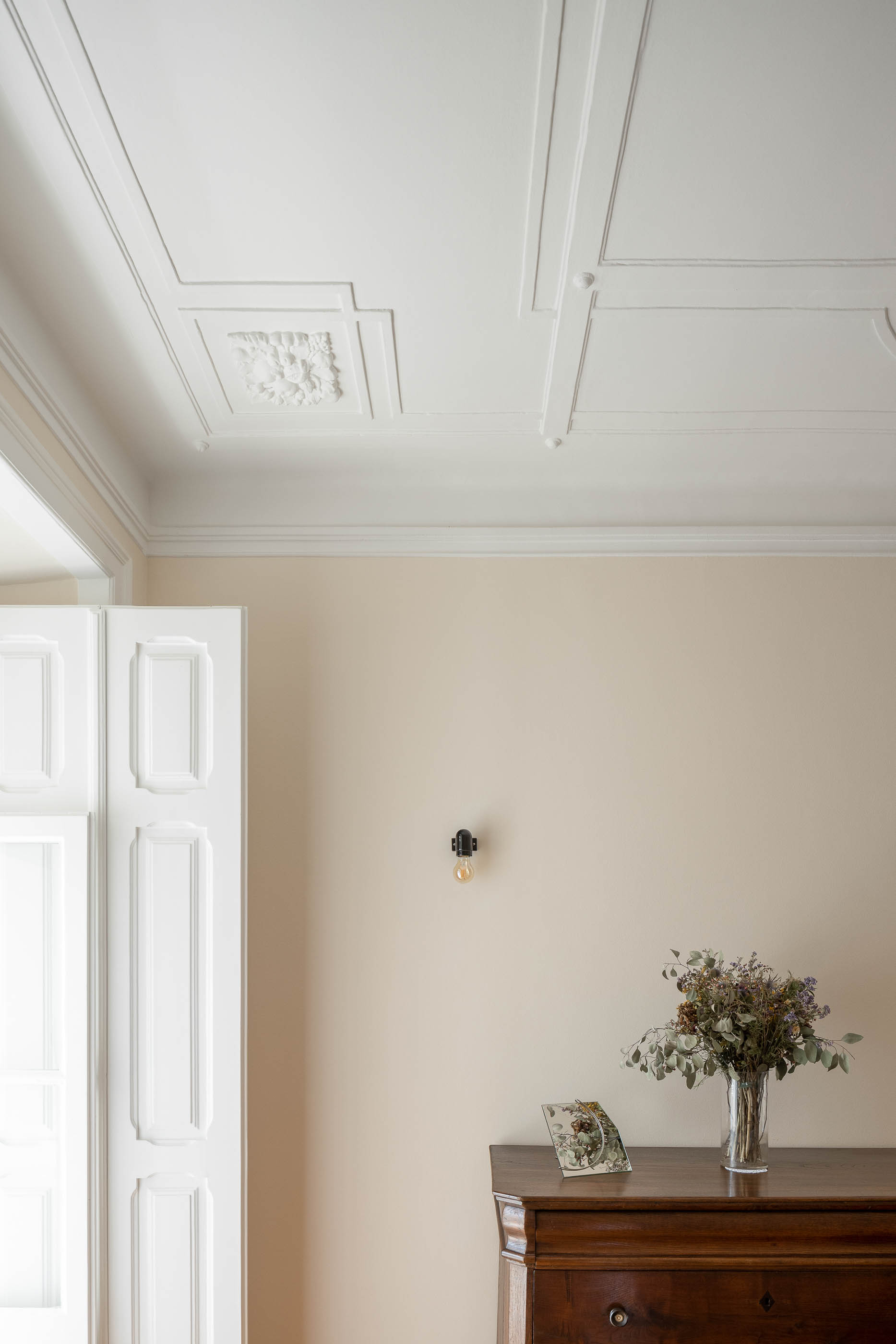 Quarto: Tecto trabalhado, cómoda antiga, candeeiro de parede, porta para varanda e casa de banho. — Apartamento da Penha arquitetura Paulo Moreira Architectures em Lisboa