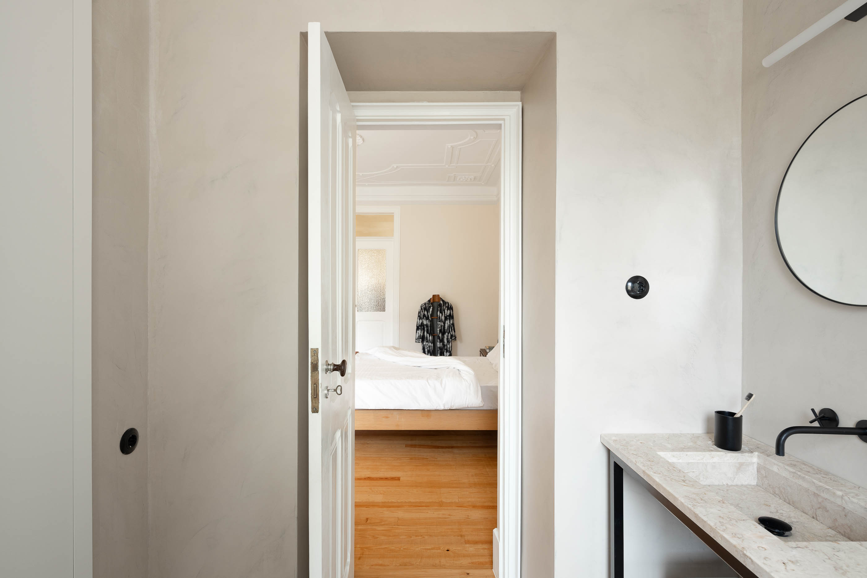Casa de banho: Porta aberta para quarto com cama de madeira, lavatório em pedra, espelho redondo. — Apartamento da Penha arquitetura Paulo Moreira Architectures em Lisboa