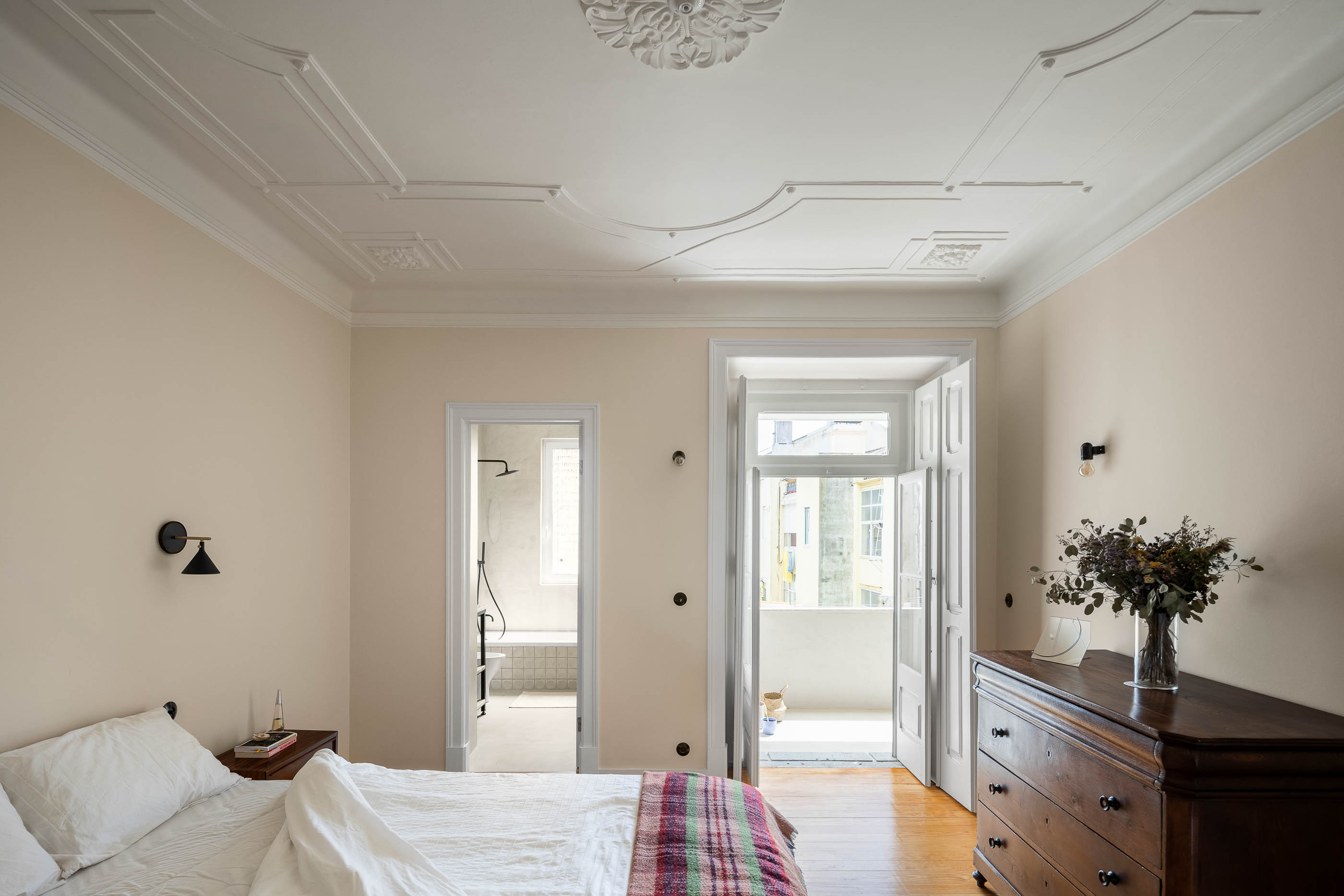 Quarto: Cama com roupa de cama branca e manta colorida, cómoda antiga, porta para casa de banho e varanda. — Apartamento da Penha arquitetura Paulo Moreira Architectures em Lisboa
