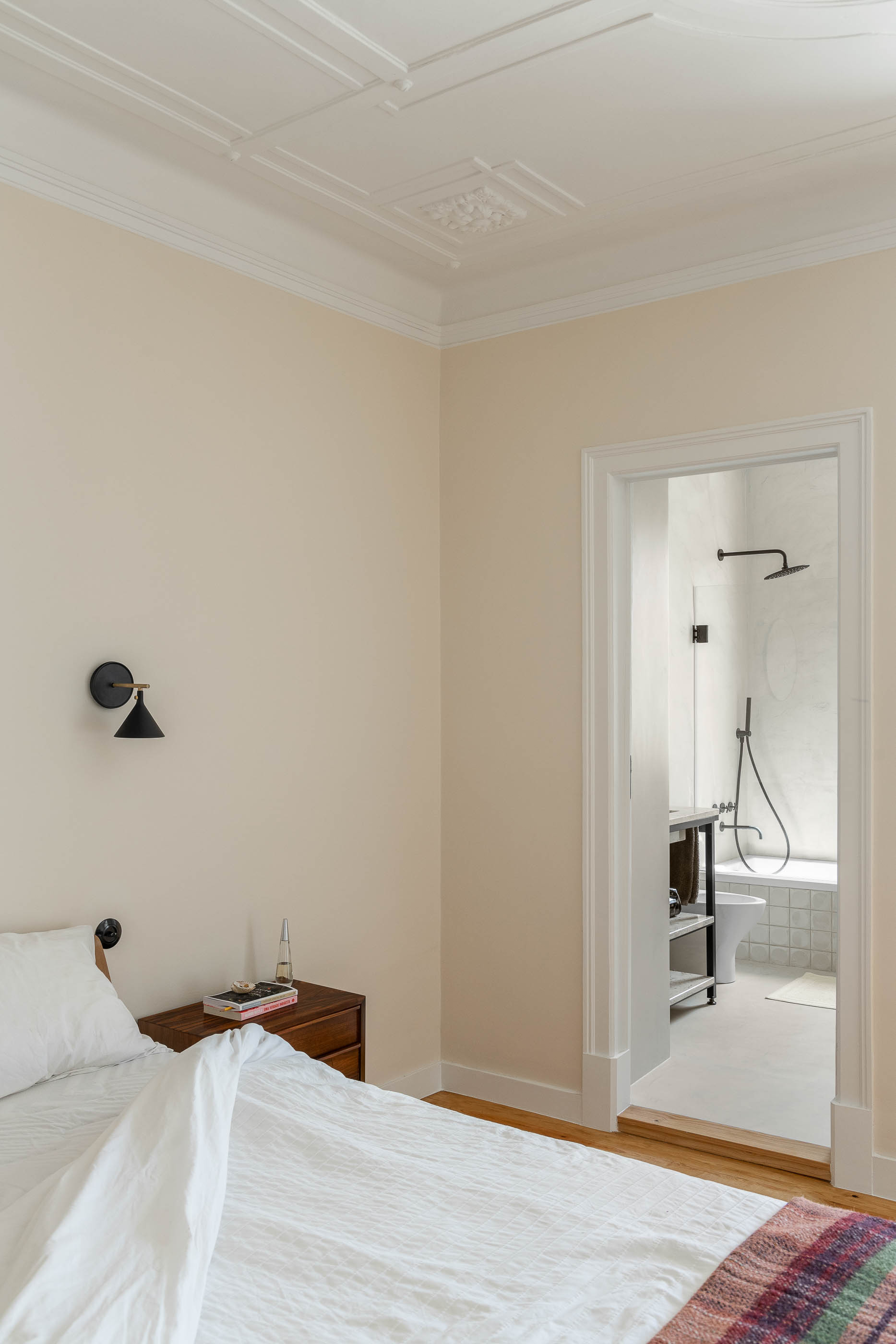 Quarto: Cama com roupa de cama branca, mesa de cabeceira em madeira, porta aberta para casa de banho. — Apartamento da Penha arquitetura Paulo Moreira Architectures em Lisboa