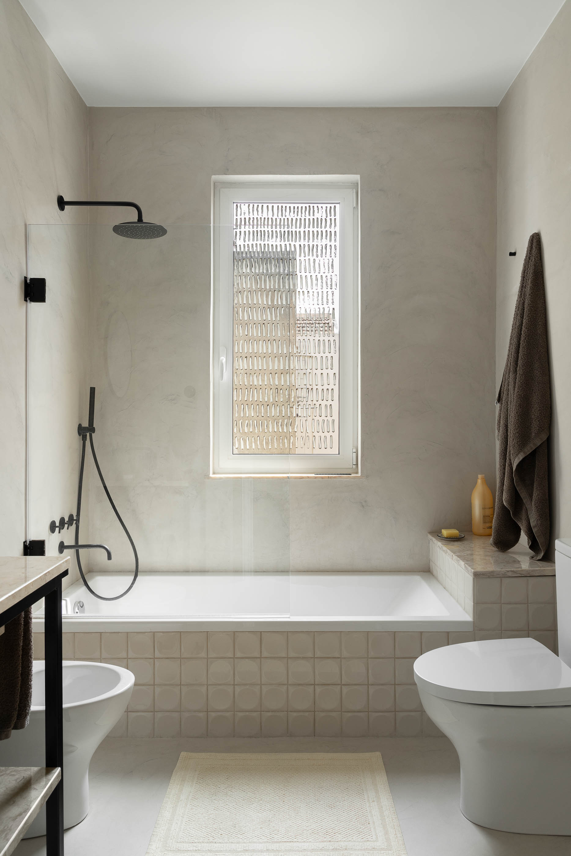 Casa de banho: Banheira e duche com resguardo em vidro, sanita e bidé, janela com padrão. — Apartamento da Penha arquitetura Paulo Moreira Architectures em Lisboa