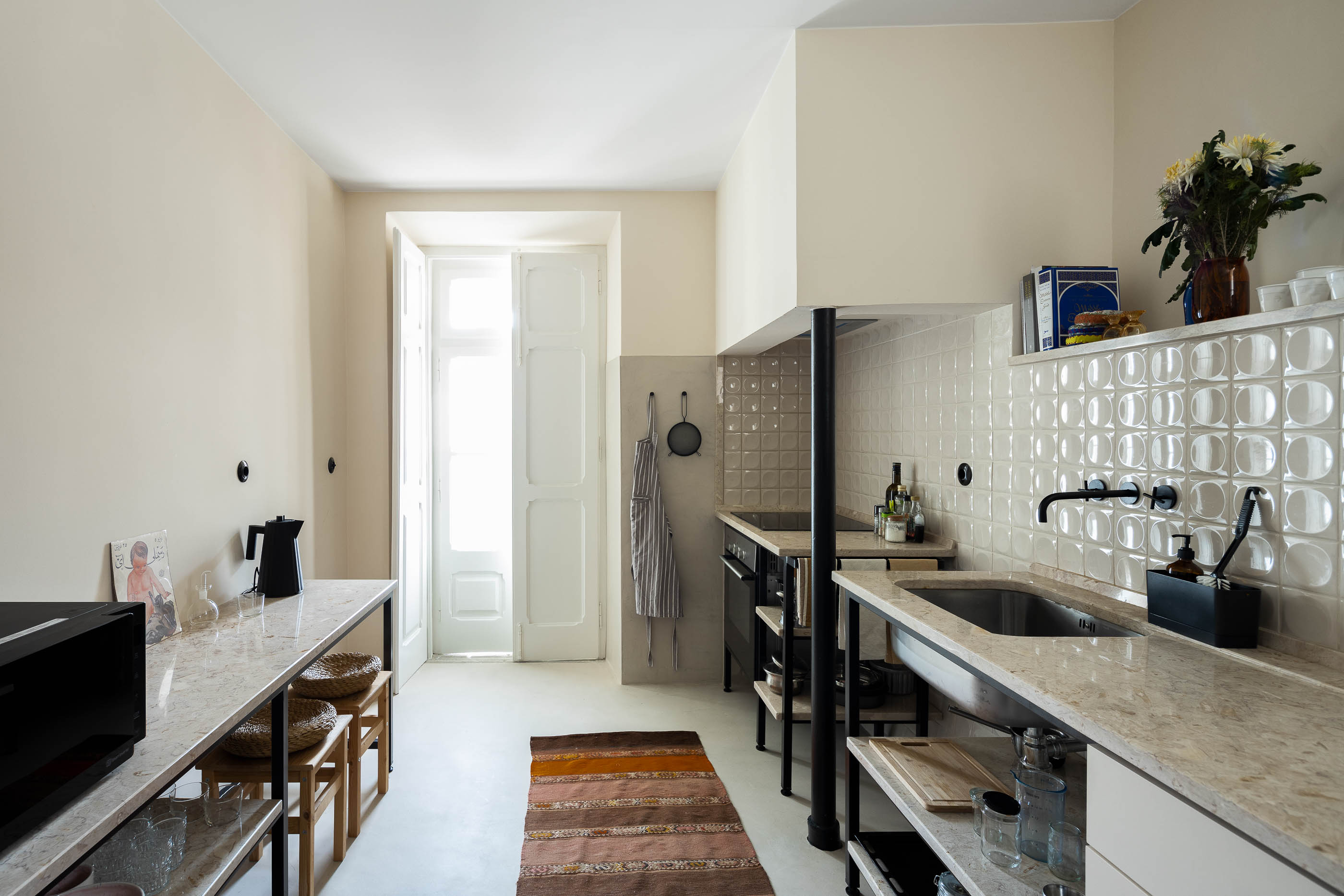 Cozinha: Bancada em pedra com estrutura metálica, mosaico branco na parede, pavimento claro, alcatifa comprida. — Apartamento da Penha arquitetura Paulo Moreira Architectures em Lisboa