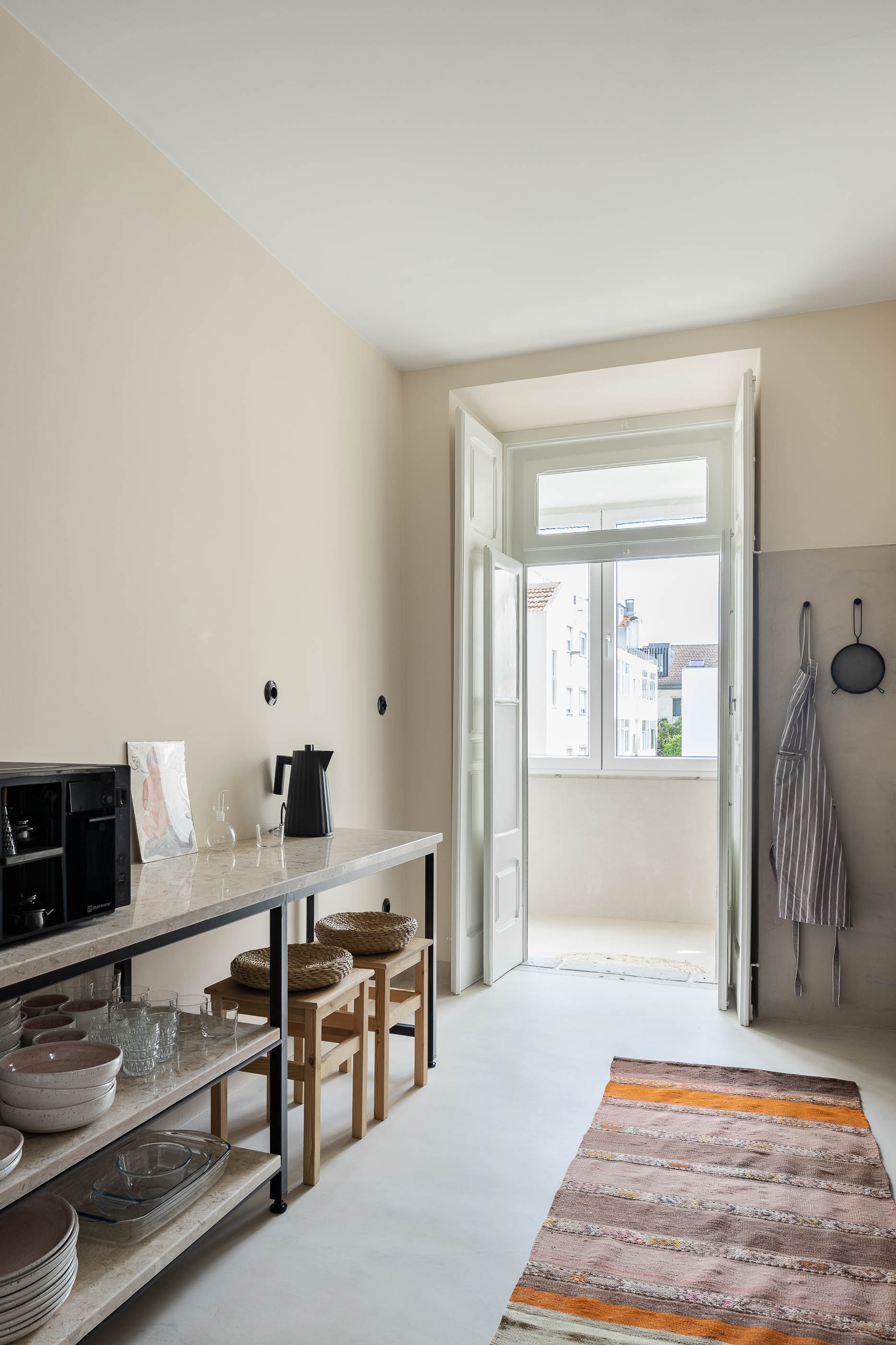 Cozinha: Bancada em pedra com estrutura metálica, pavimento claro, porta para varanda, alcatifa comprida. — Apartamento da Penha arquitetura Paulo Moreira Architectures em Lisboa