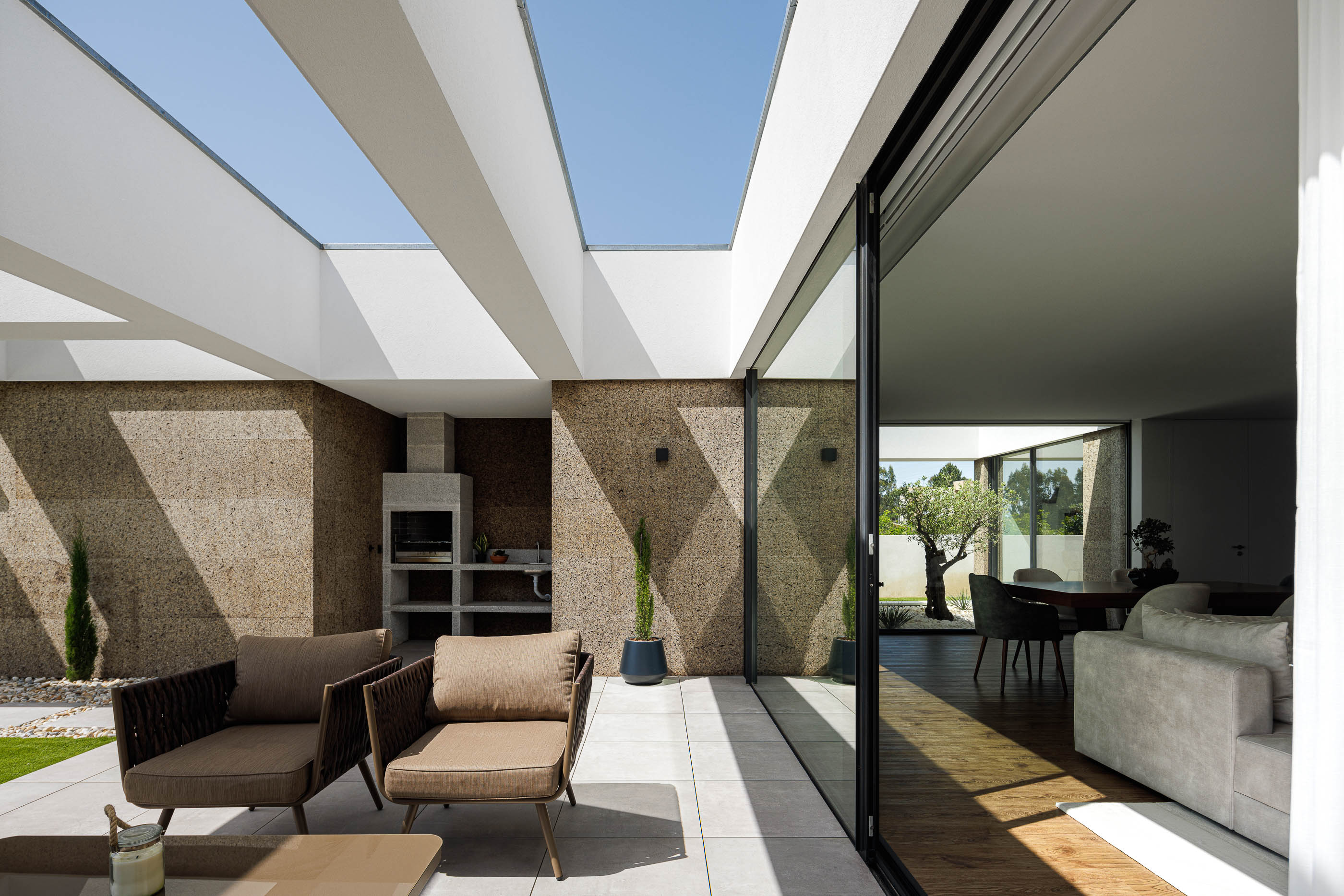 Alpendre: Zona de estar exterior com churrasqueira, pavimento cerâmico e acesso à sala através de amplas janelas. — Casa AS arquitetura Mário Alves Arquitecto em Santa Maria da Feira