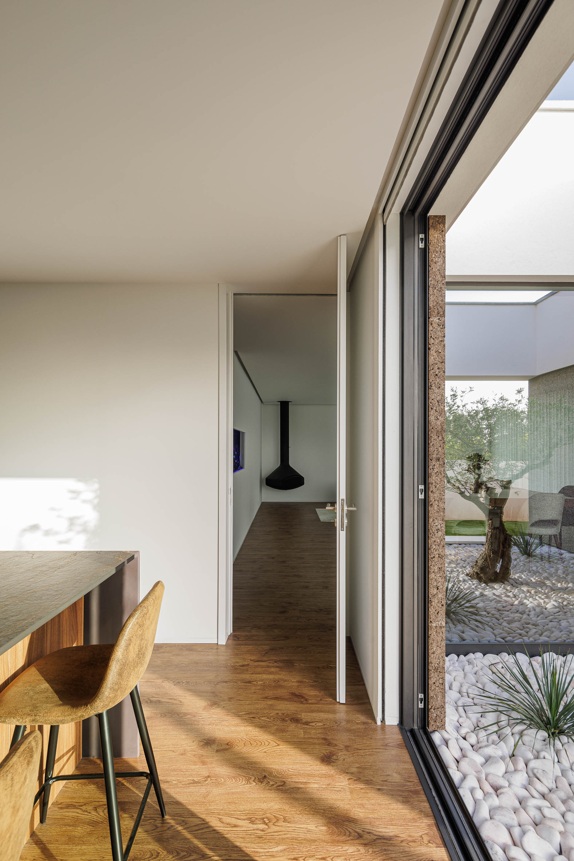 Cozinha: Vista da cozinha para o pátio interior, com ilha, cadeiras altas e porta para outra divisão. — Casa AS arquitetura Mário Alves Arquitecto em Santa Maria da Feira
