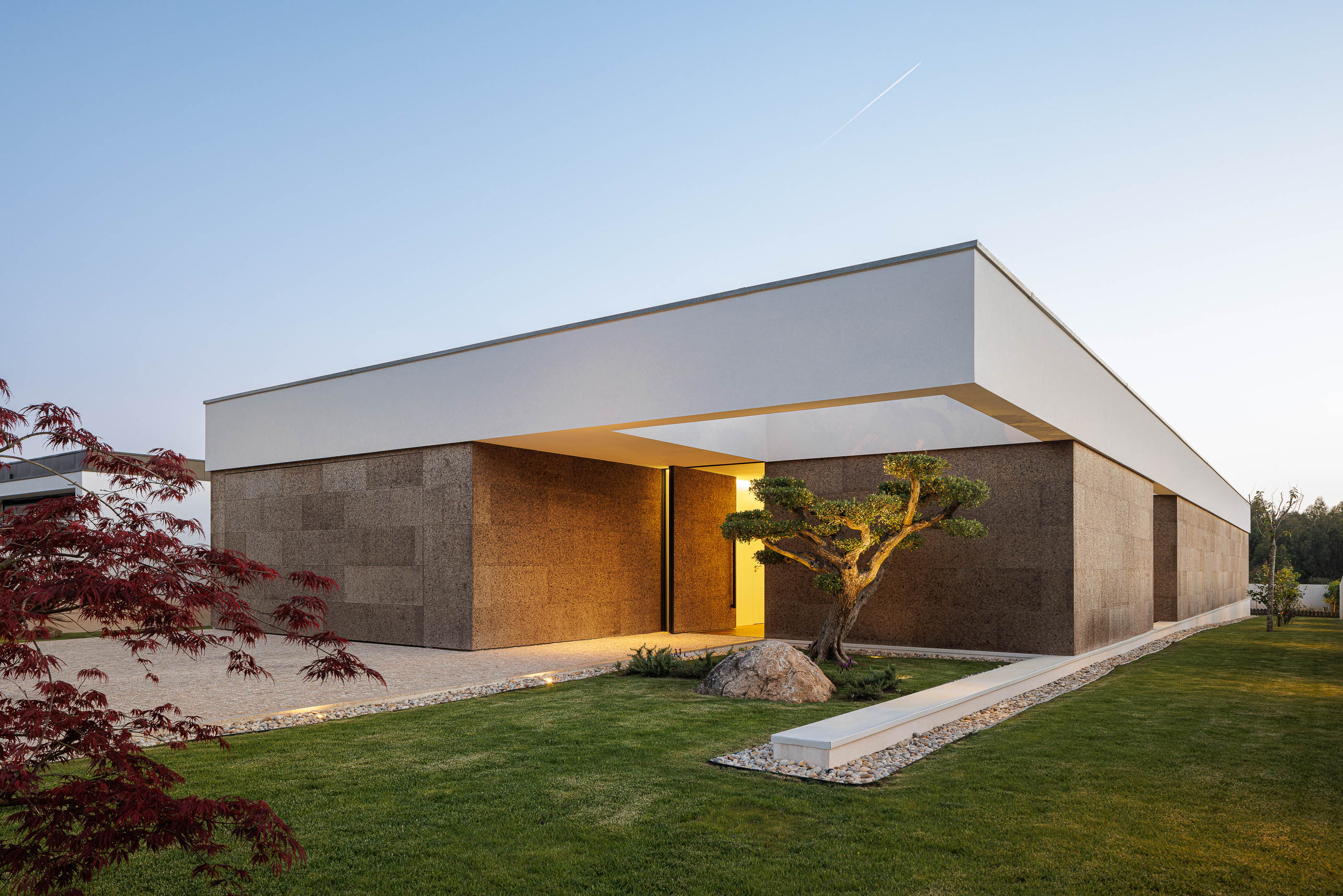 Fachada: Moradia térrea com revestimento em cortiça, volume branco superior e jardim com árvore de folha vermelha. — Casa AS arquitetura Mário Alves Arquitecto em Santa Maria da Feira