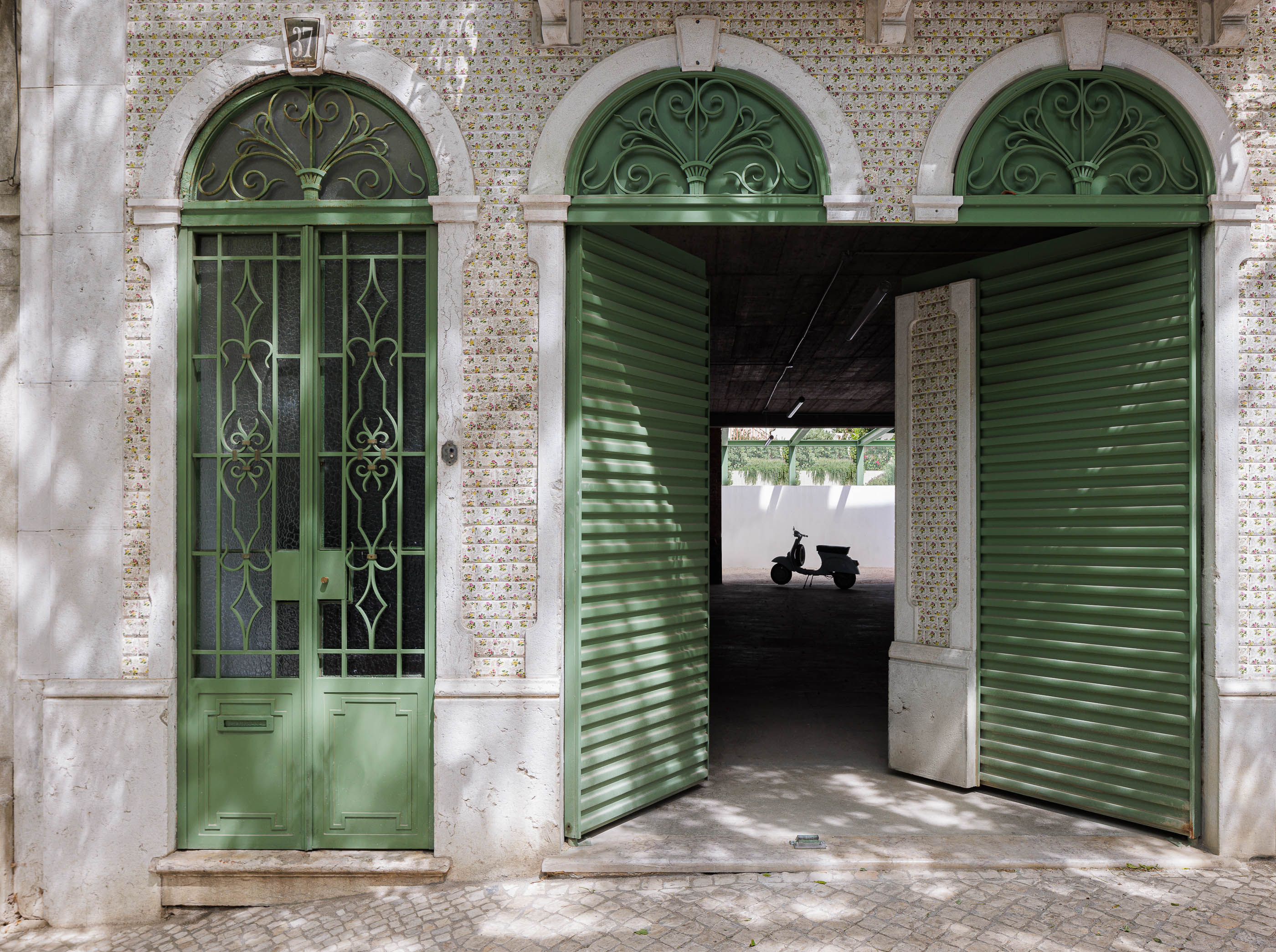Garagem: Entrada com portões verdes abertos, revelando interior amplo e uma mota clássica. — Possidónio da Silva 37 arquitetura Fragmentos em Lisboa