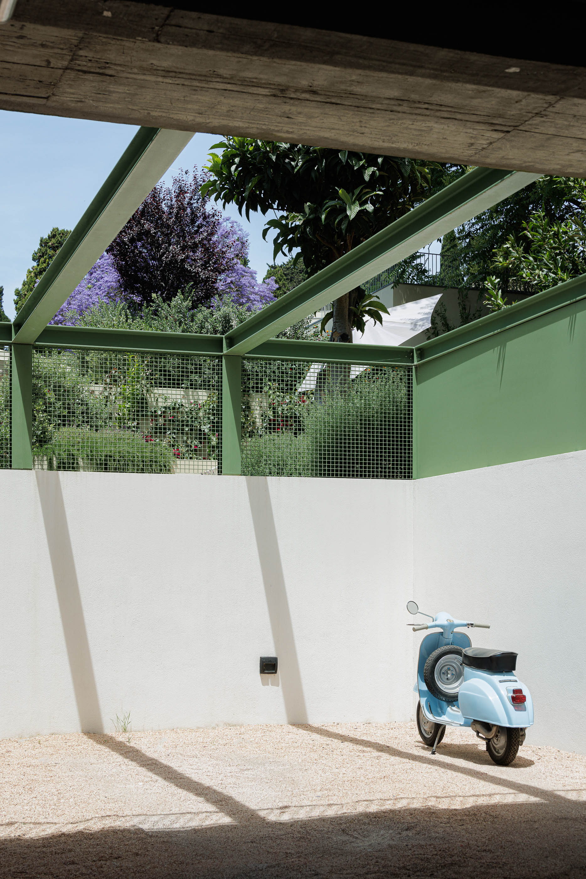 Garagem: Mota azul clara estacionada num pátio com pavimento de gravilha e estrutura metálica verde. — Possidónio da Silva 37 arquitetura Fragmentos em Lisboa