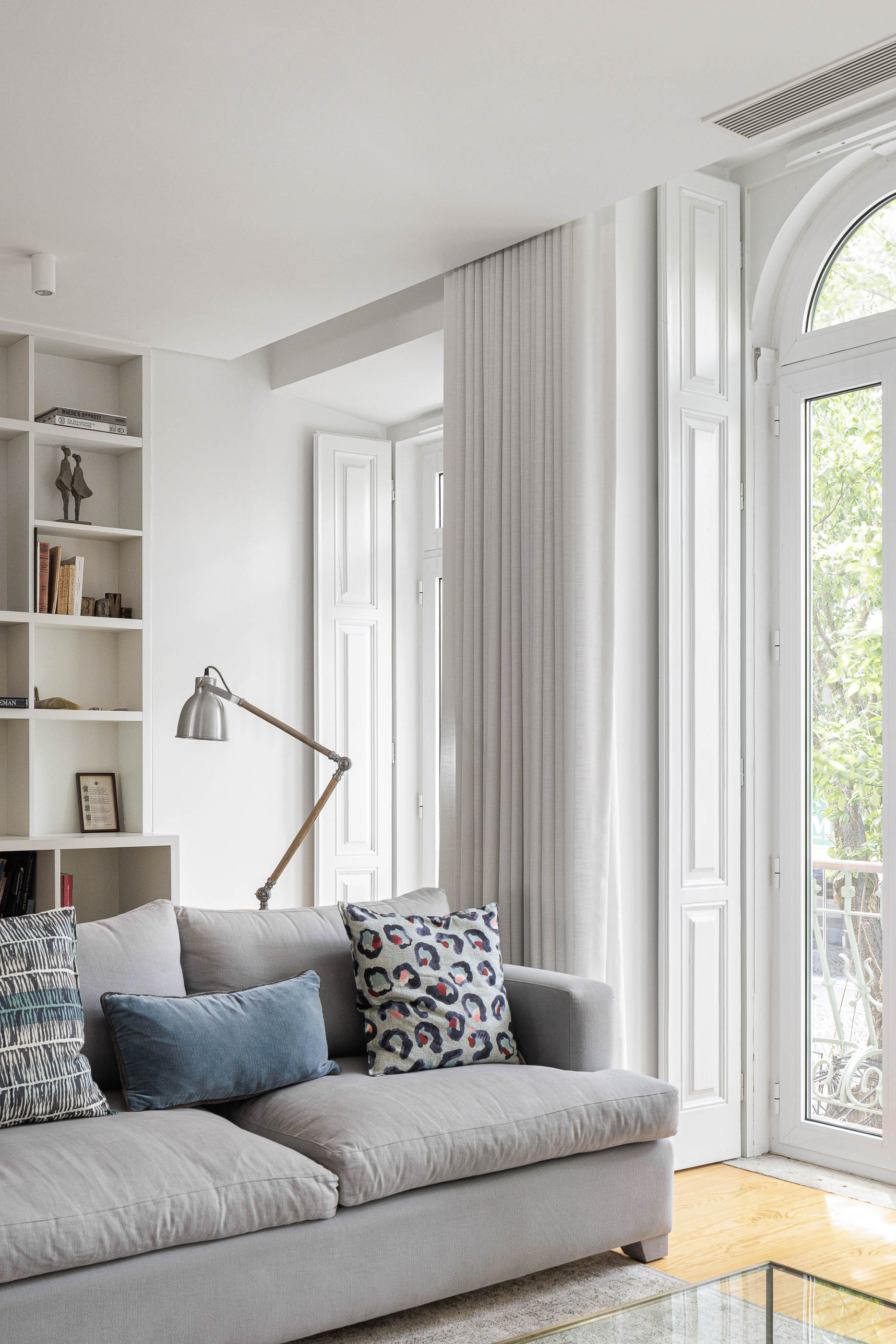 Sala de estar: Sofá cinzento com almofadas estampadas, candeeiro de leitura e janela com portadas brancas. — Possidónio da Silva 37 arquitetura Fragmentos em Lisboa