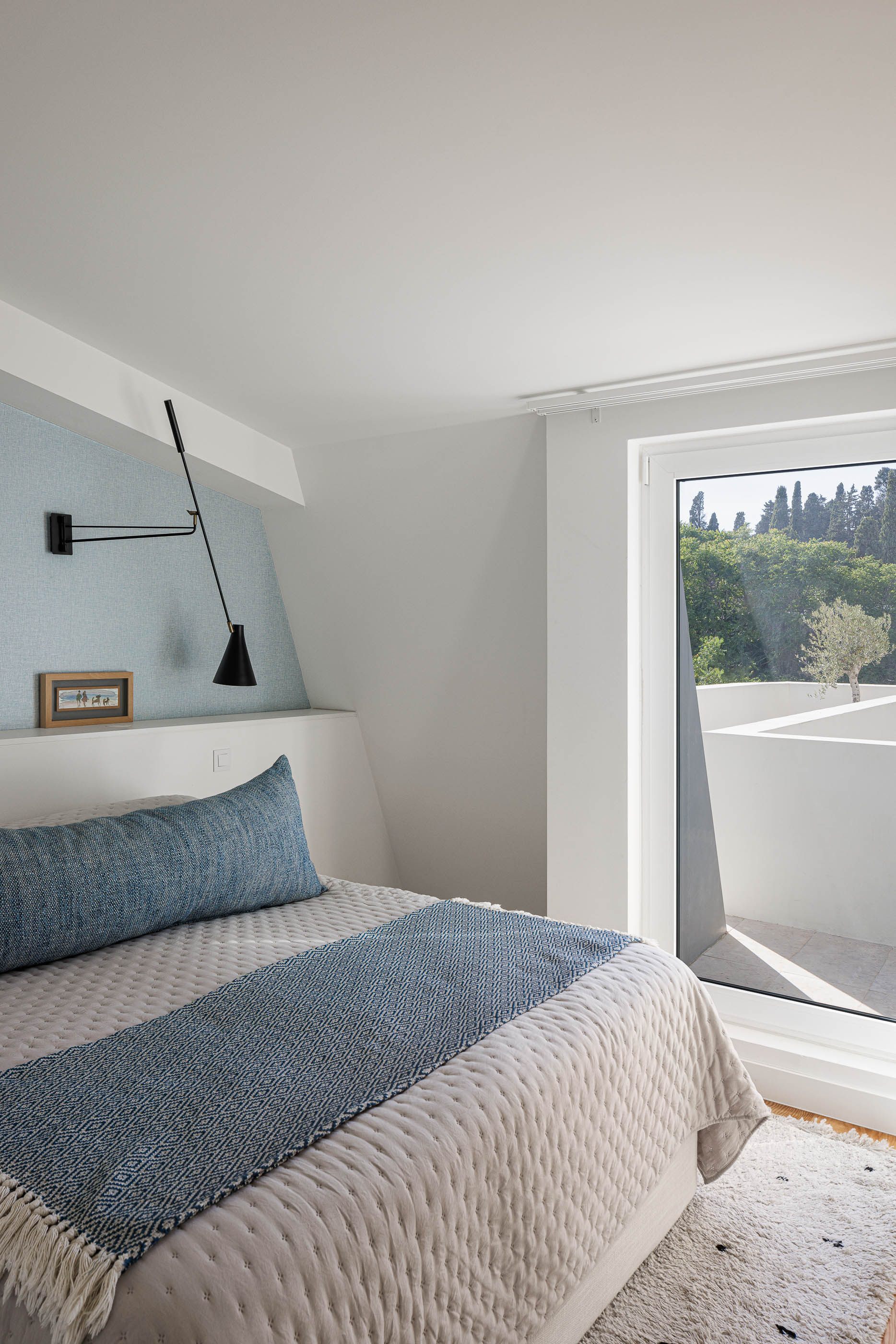 Quarto: Cama com cabeceira azul, candeeiro de parede preto e acesso a terraço com vista para árvores. — Possidónio da Silva 37 arquitetura Fragmentos em Lisboa