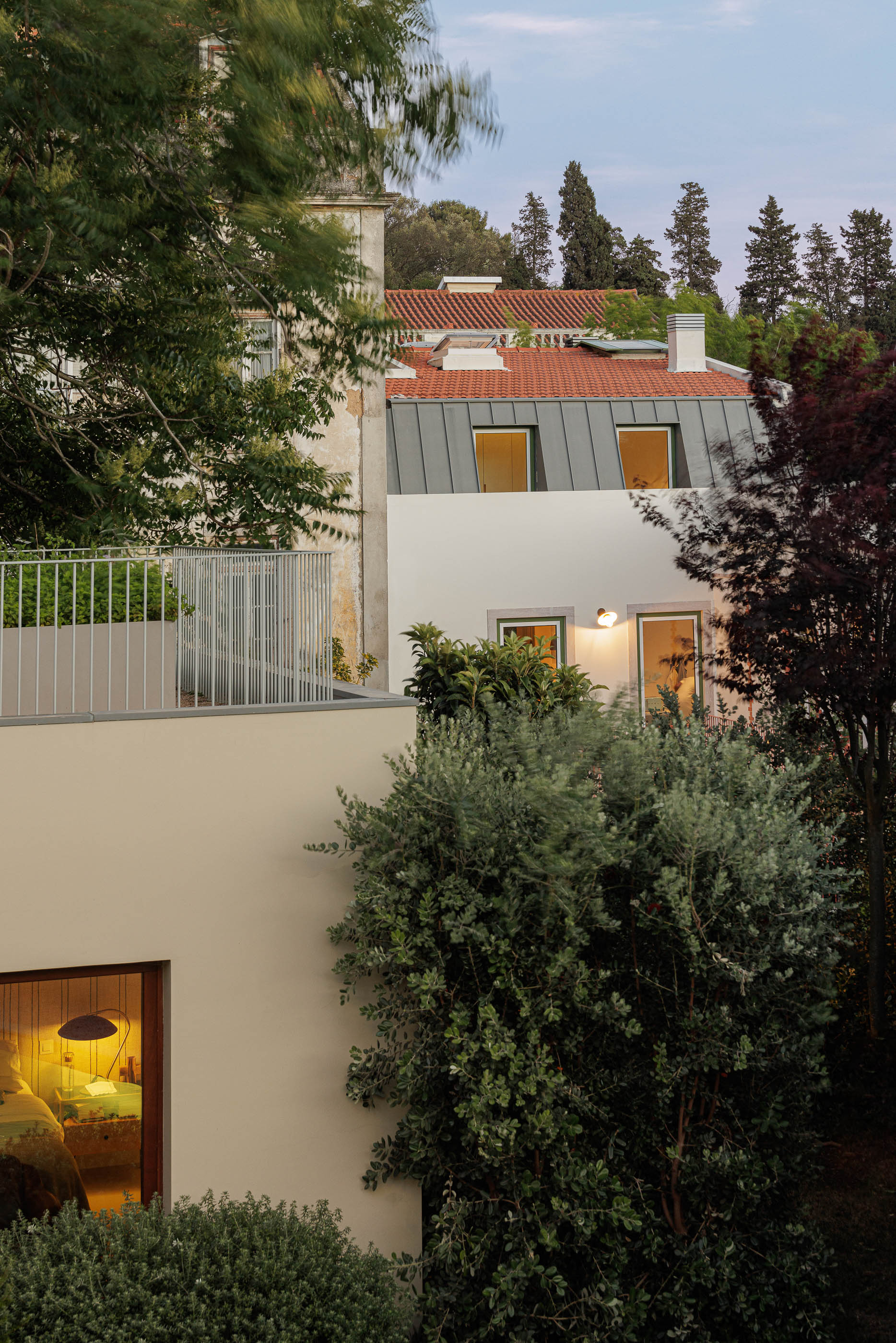 Exterior: Vista noturna da casa com telhado cinzento, candeeiros acesos e vegetação. — Possidónio da Silva 37 arquitetura Fragmentos em Lisboa