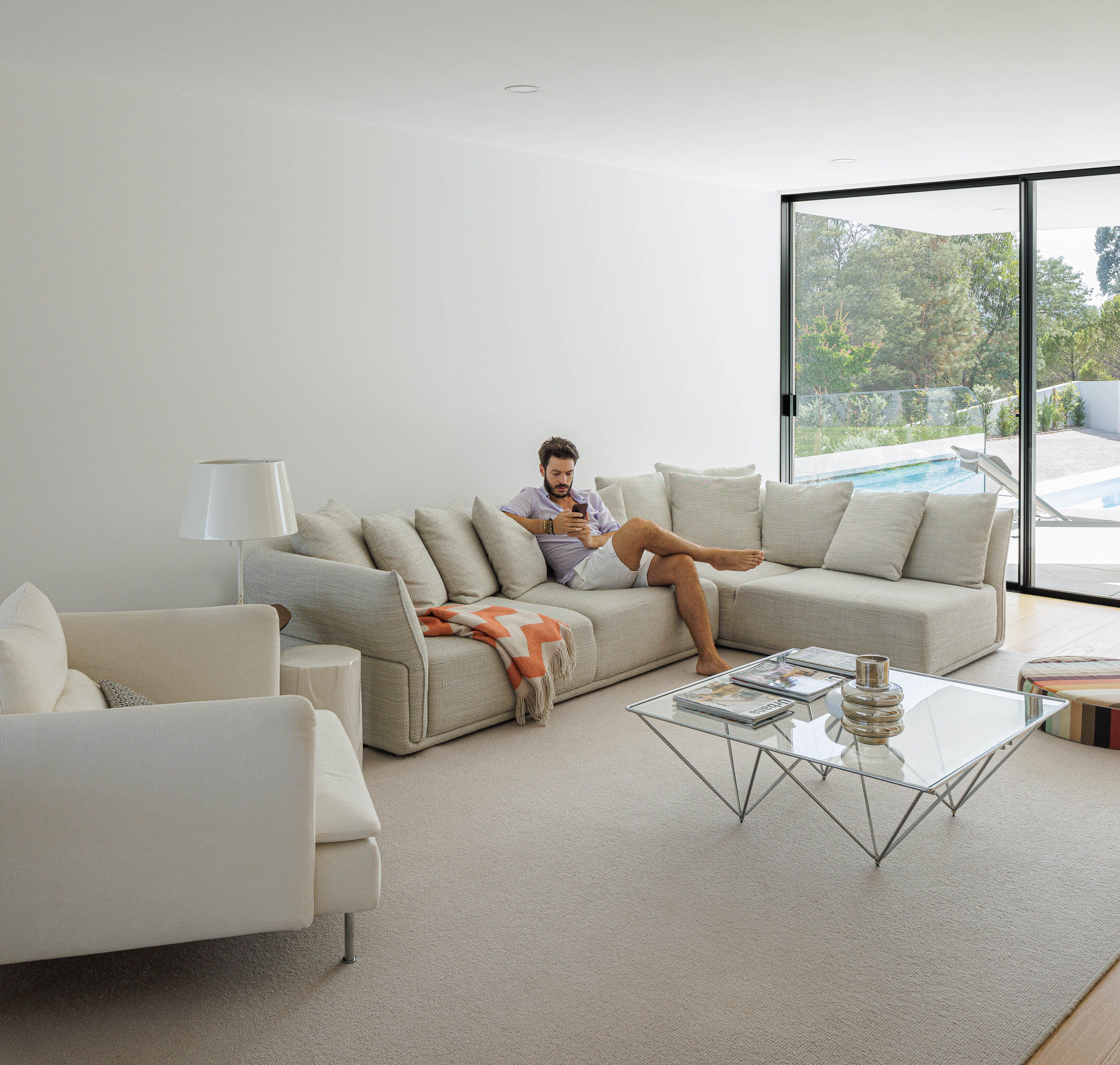 Sala de estar com sofá modular cinza, alcatifa bege e mesa de centro de vidro. Grande janela com vista para a piscina. — Casa JC arquitetura Mário Alves Arquitecto em Coimbra