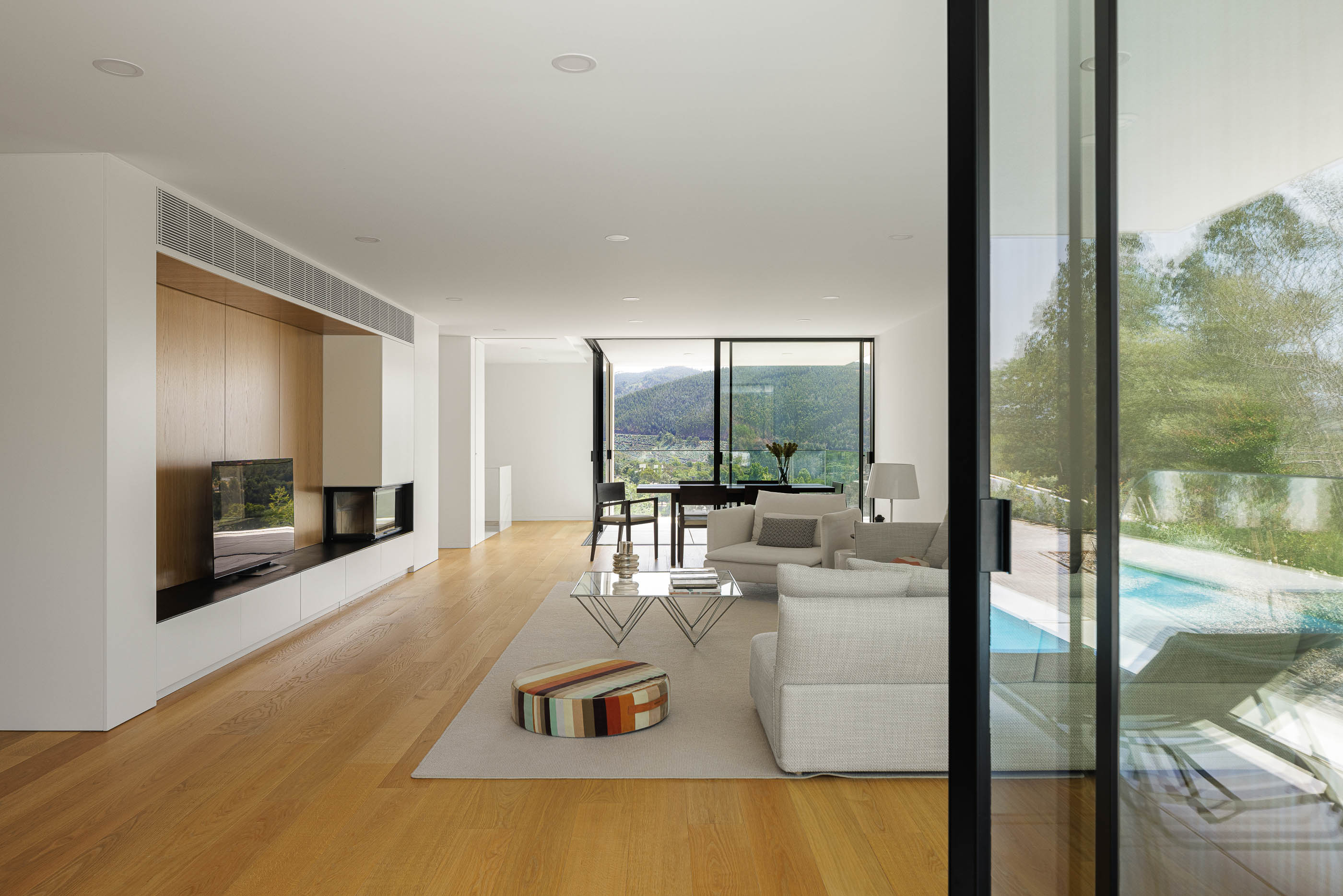Sala de estar com vista para a piscina. Pavimento em madeira clara, alcatifa bege e mobiliário minimalista. — Casa JC arquitetura Mário Alves Arquitecto em Coimbra