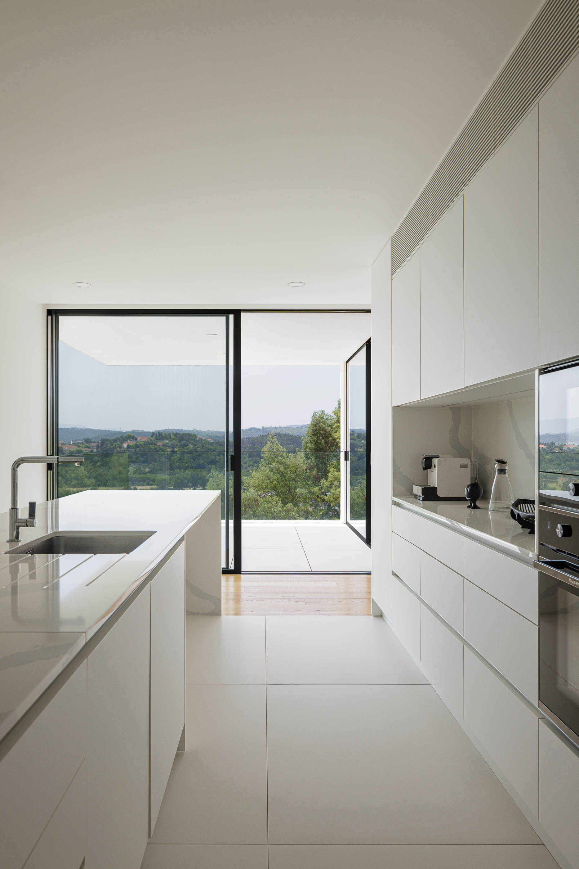 Cozinha minimalista com armários brancos e bancada em pedra clara. Pavimento cerâmico branco e vista para a paisagem. — Casa JC arquitetura Mário Alves Arquitecto em Coimbra