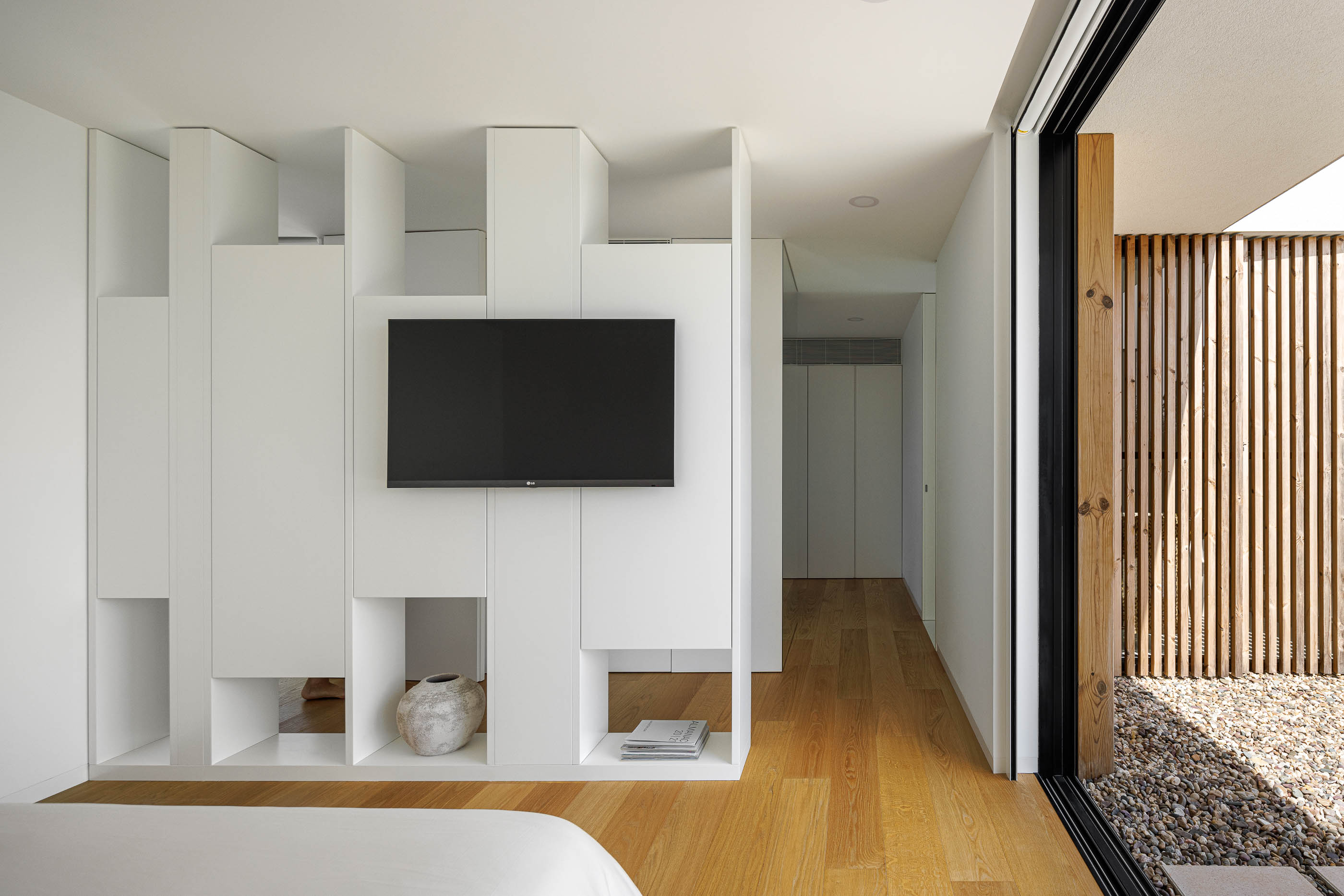 Quarto com cama, móvel branco com televisão e pavimento em madeira clara. Acesso ao pátio exterior com seixos. — Casa JC arquitetura Mário Alves Arquitecto em Coimbra