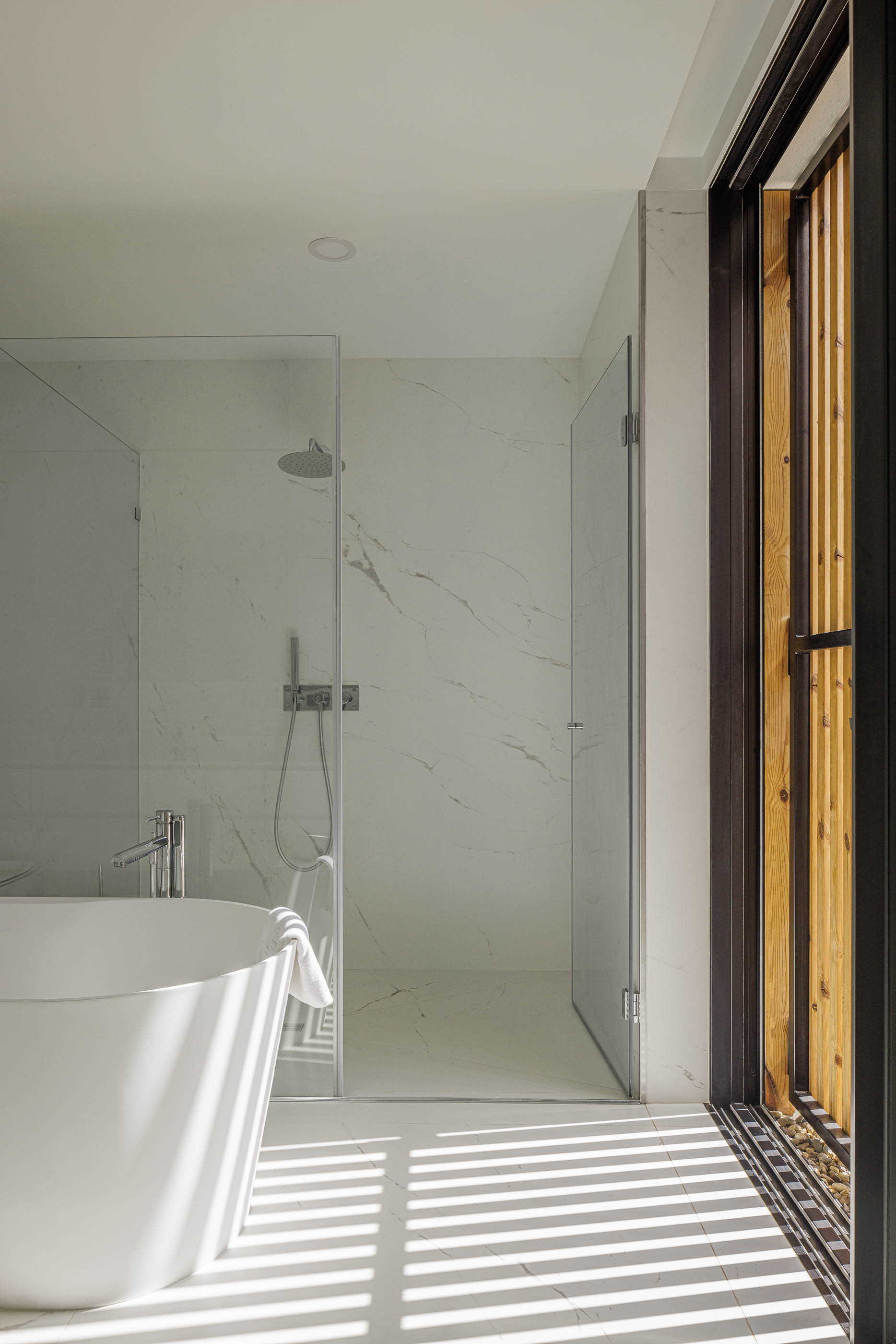 Casa de banho com banheira branca e cabine de duche em vidro. Revestimento em mármore branco e pavimento cerâmico. — Casa JC arquitetura Mário Alves Arquitecto em Coimbra
