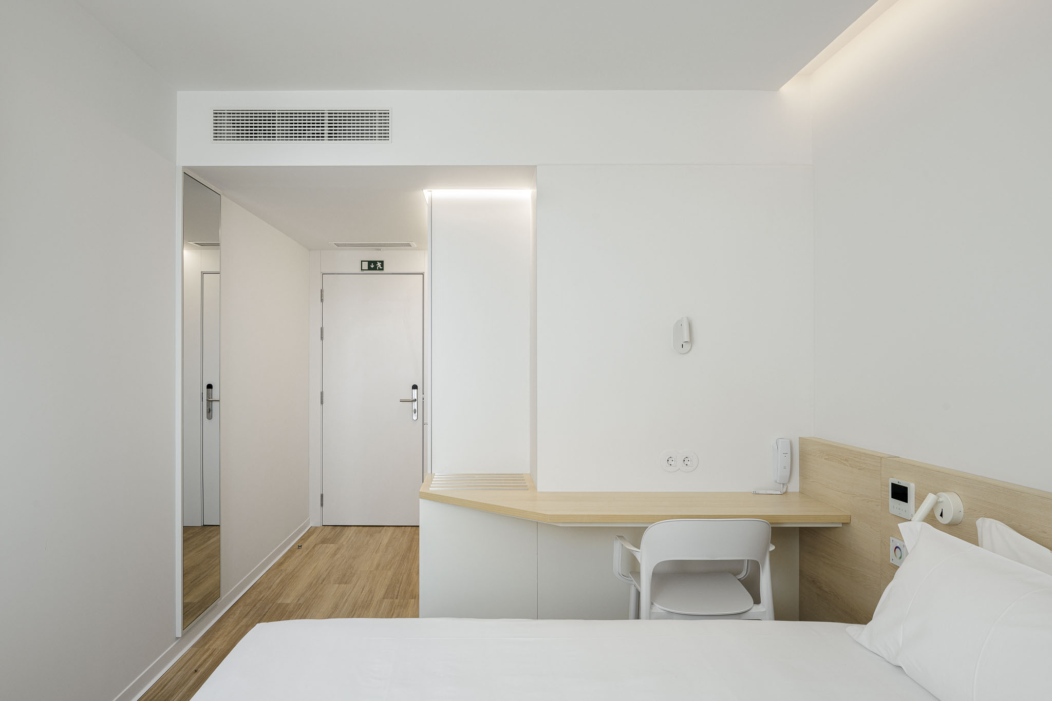 Quarto: Quarto branco com secretária de madeira clara, cadeira branca e roupeiro com espelho. — Hotel B&B Famalicão arquitetura Jorge Domingues Arquitetos em Famalicão
