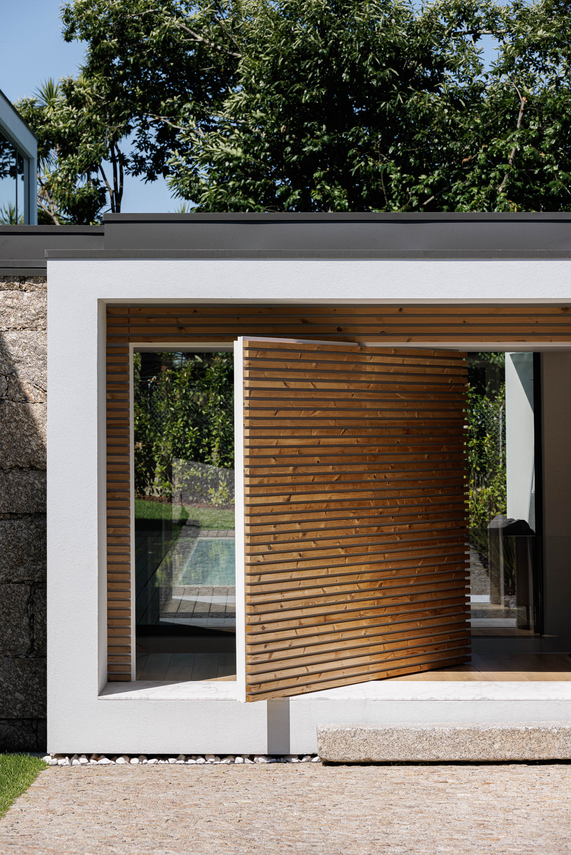 Entrada: Porta pivotante em madeira ripada, embutida em estrutura branca, pavimento exterior em calçada. — CASA [EN]COSTA arquitetura Hous3 em Guimarães