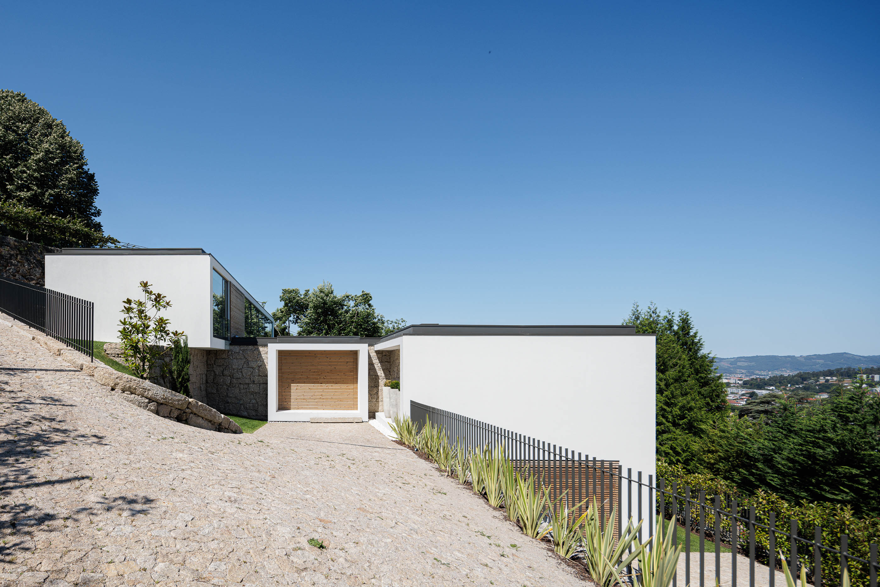 Exterior: Fachada minimalista branca com detalhes em madeira, integrada na encosta com muros de pedra. — CASA [EN]COSTA arquitetura Hous3 em Guimarães