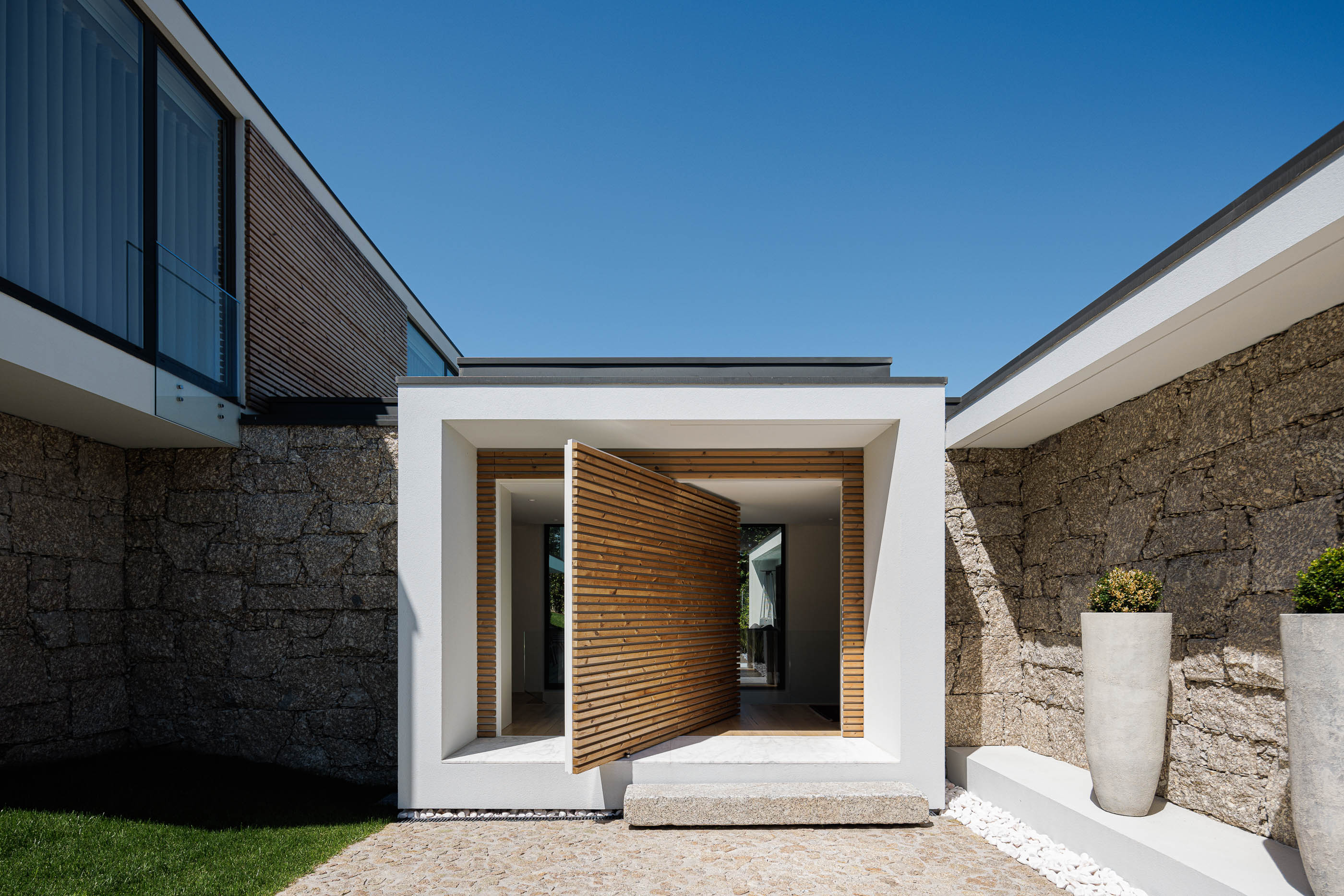 Entrada: Perspectiva frontal da entrada principal com porta pivotante em madeira e paredes em pedra. — CASA [EN]COSTA arquitetura Hous3 em Guimarães